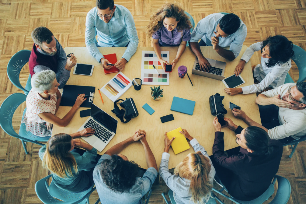 Overhead view of diverse team collaborating around table with laptops, tablets, and design materials during group meeting.