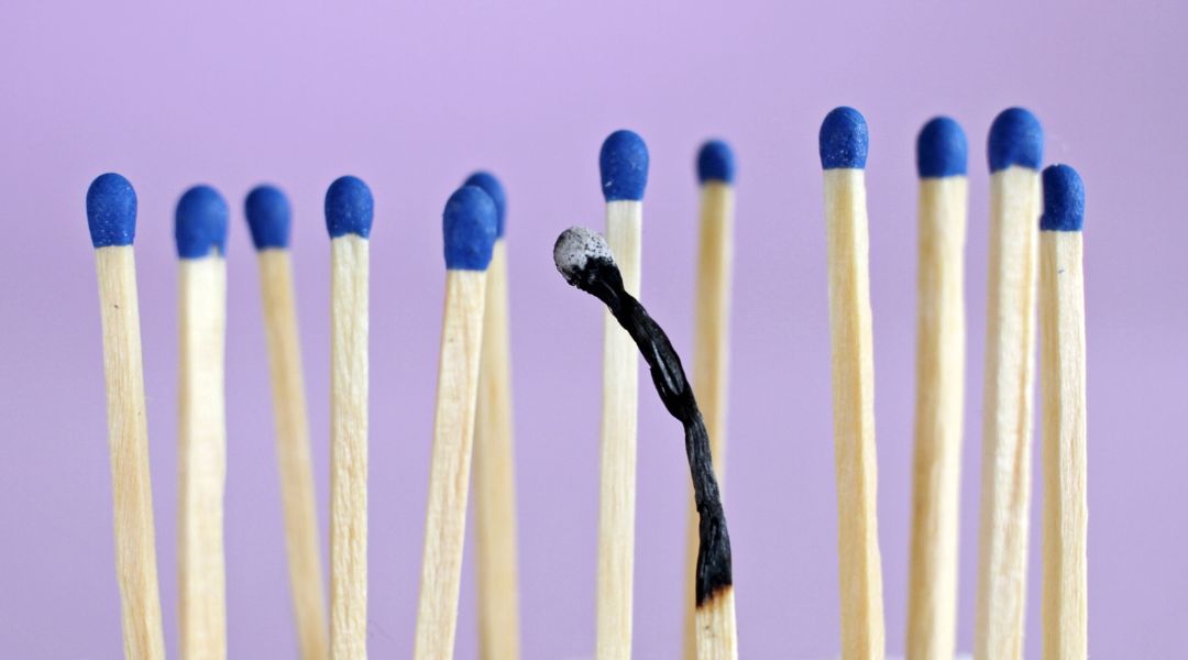 Single burnt matchstick surrounded by unlit blue-tipped matches on a purple background, symbolizing burnout.
