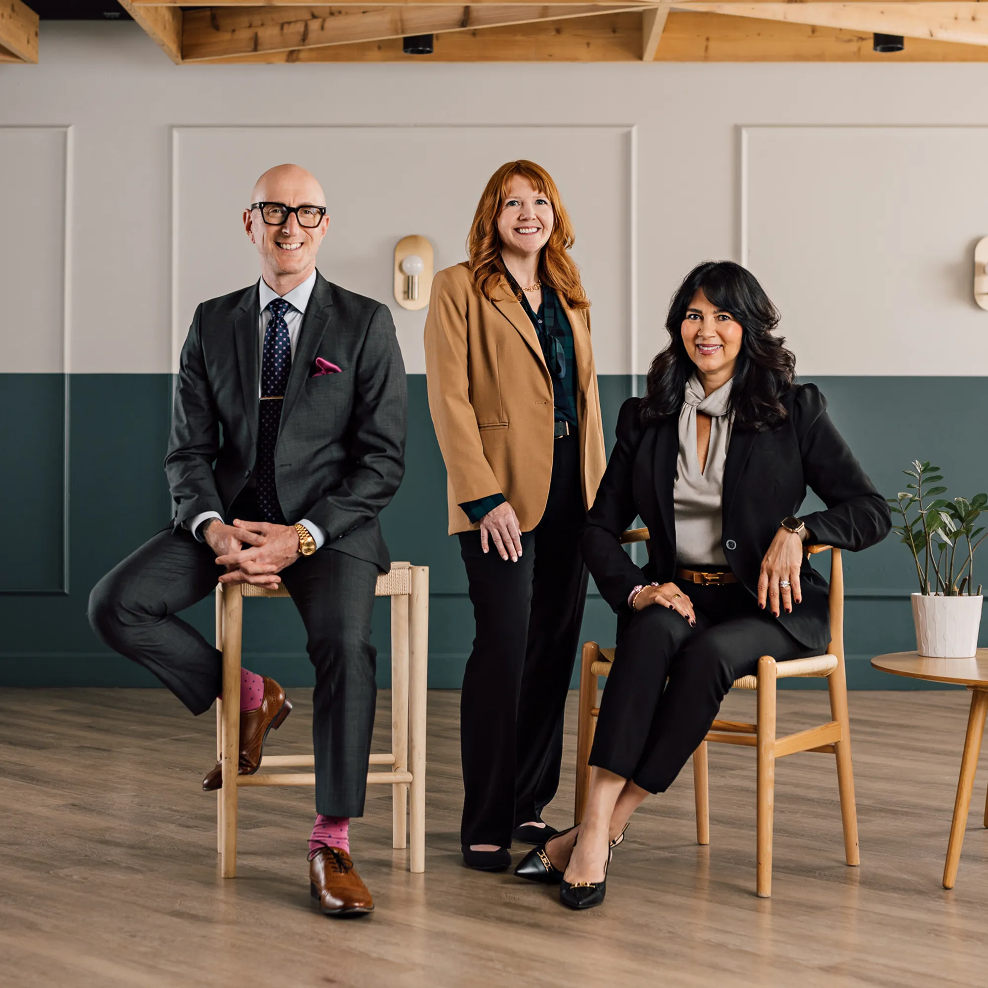 Aram Arslanian, Michelle Rakshys, and Michelle Winklaar pose together in a bright studio, two seated on wooden chairs and one standing between them