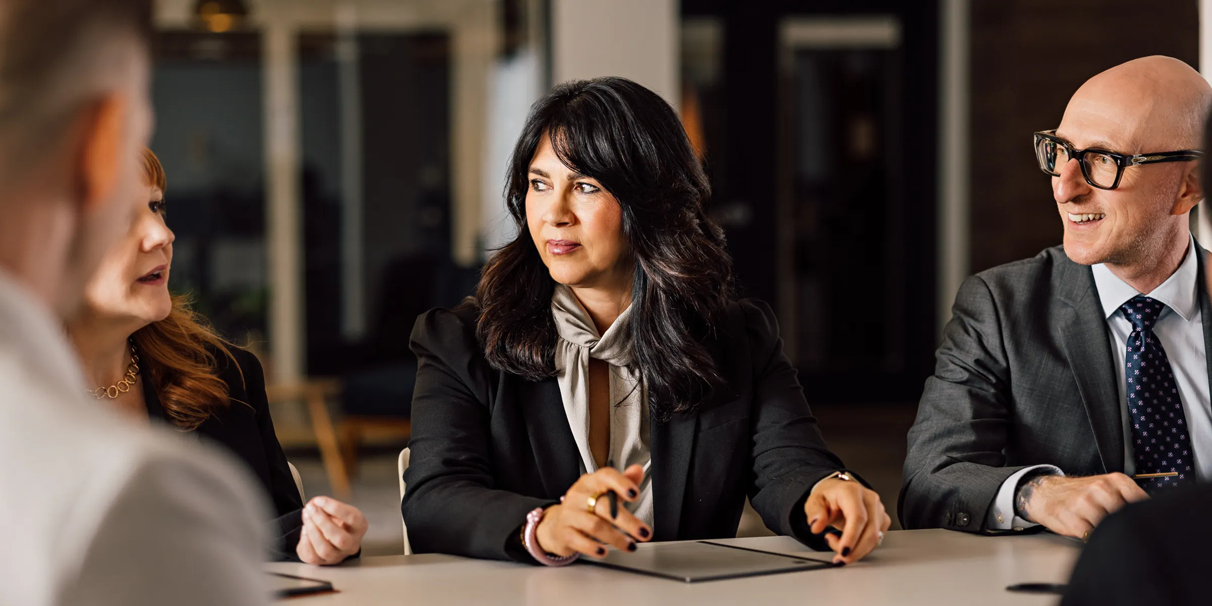Colleagues in business attire talk around a boardroom table, a woman in a black blazer at the centre