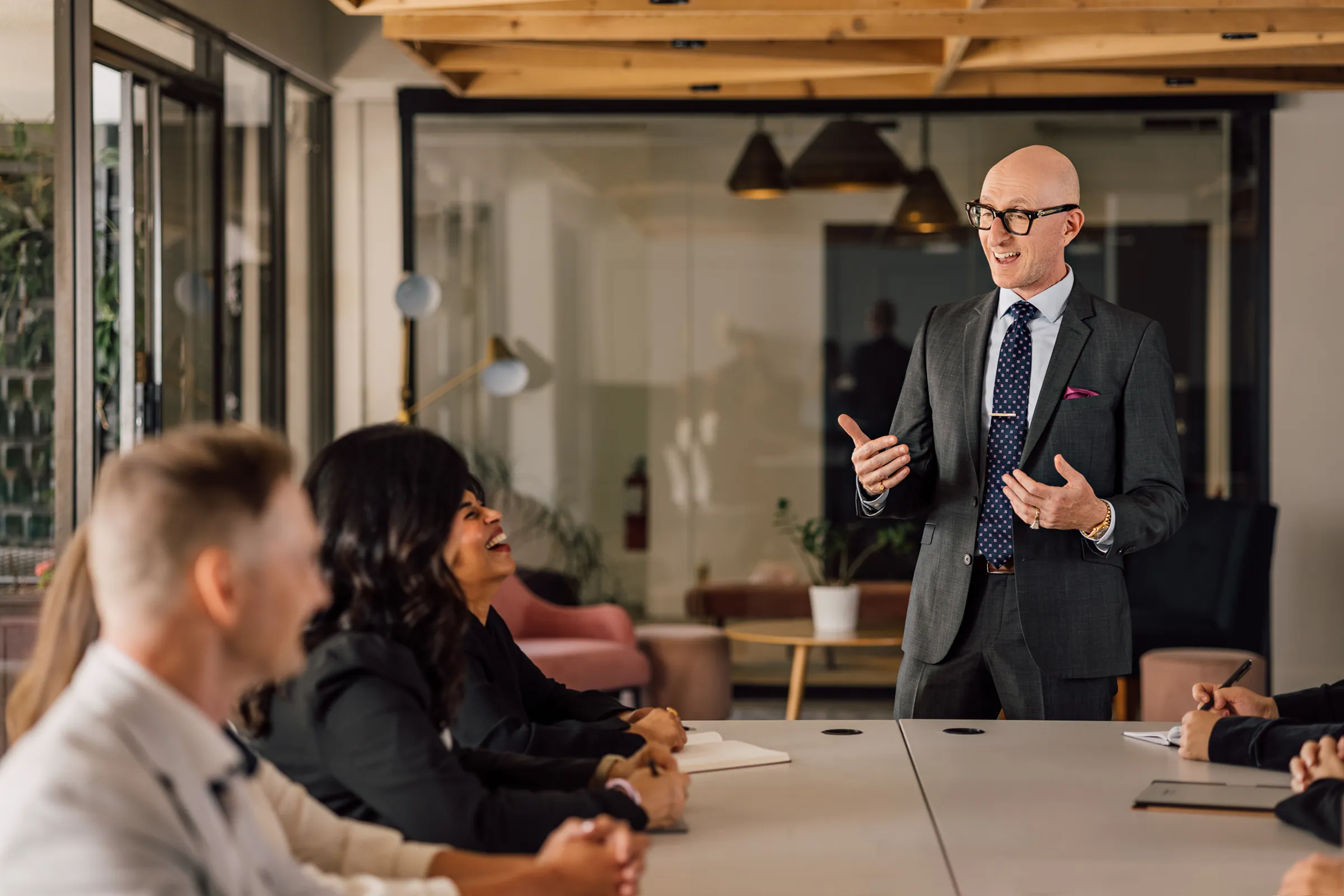 A man in a dark suit and glasses is standing and speaking to a group of people seated at a conference table in a modern office, with one woman laughing and others attentively listening.