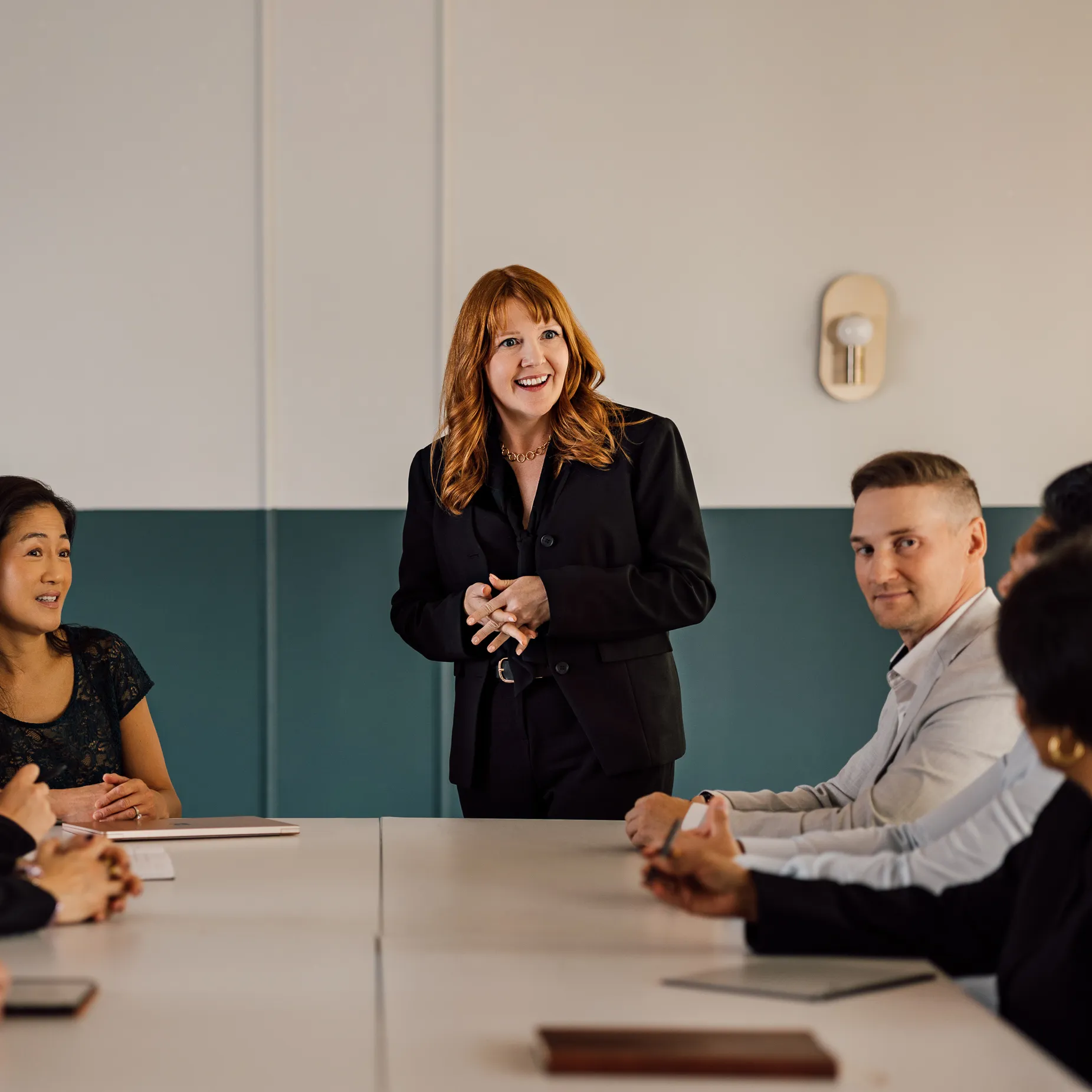 A red-haired woman in a black suit standing and speaking to a group seated around a conference table in a meeting room.