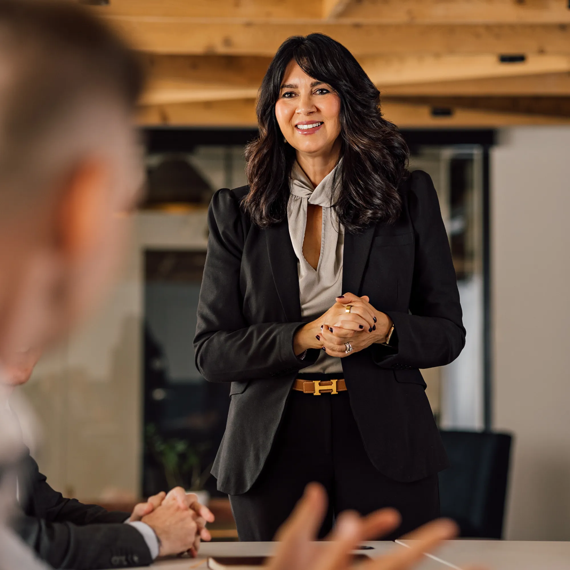Smiling businesswoman with dark wavy hair in a black blazer and beige blouse speaking in a modern office to colleagues whose blurred hands and heads are visible.