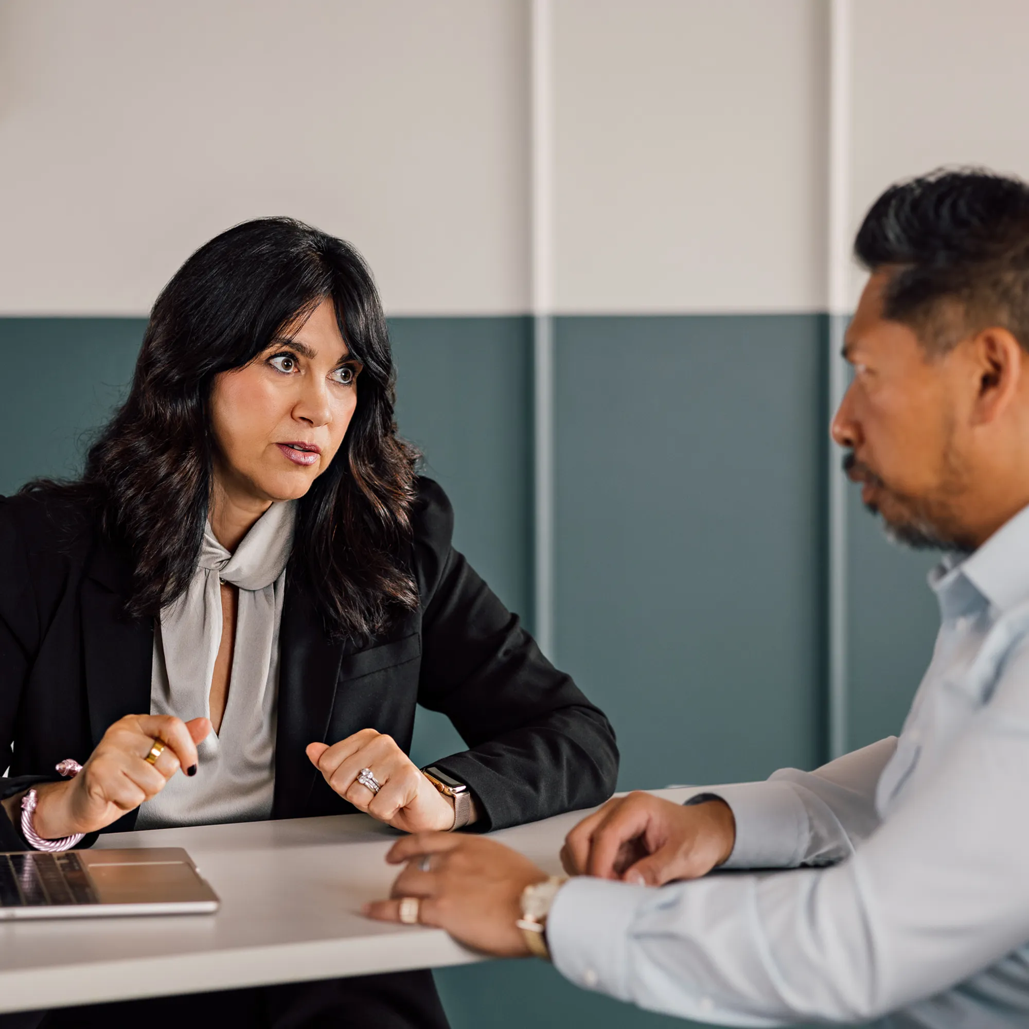 Two business professionals engaged in a serious conversation across a desk, with a laptop nearby.