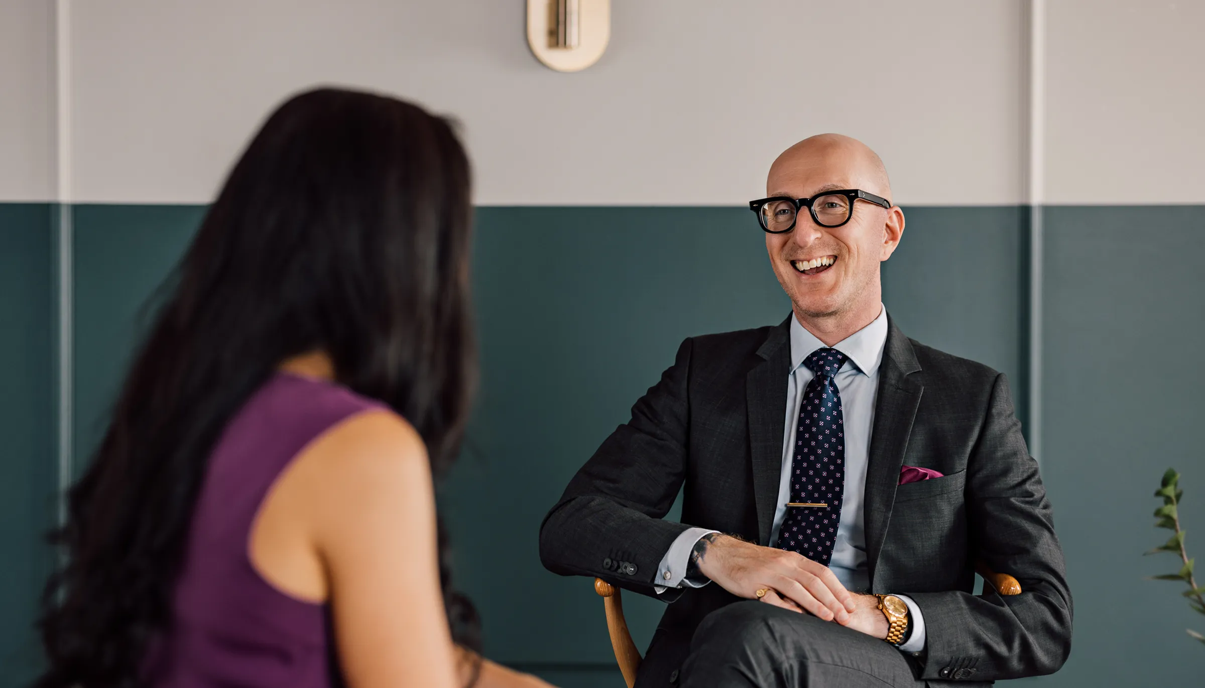 A man in glasses wearing a dark suit with a patterned tie smiles while talking to a woman with long dark hair in a sleeveless purple top, seated in a modern office with teal and white walls.