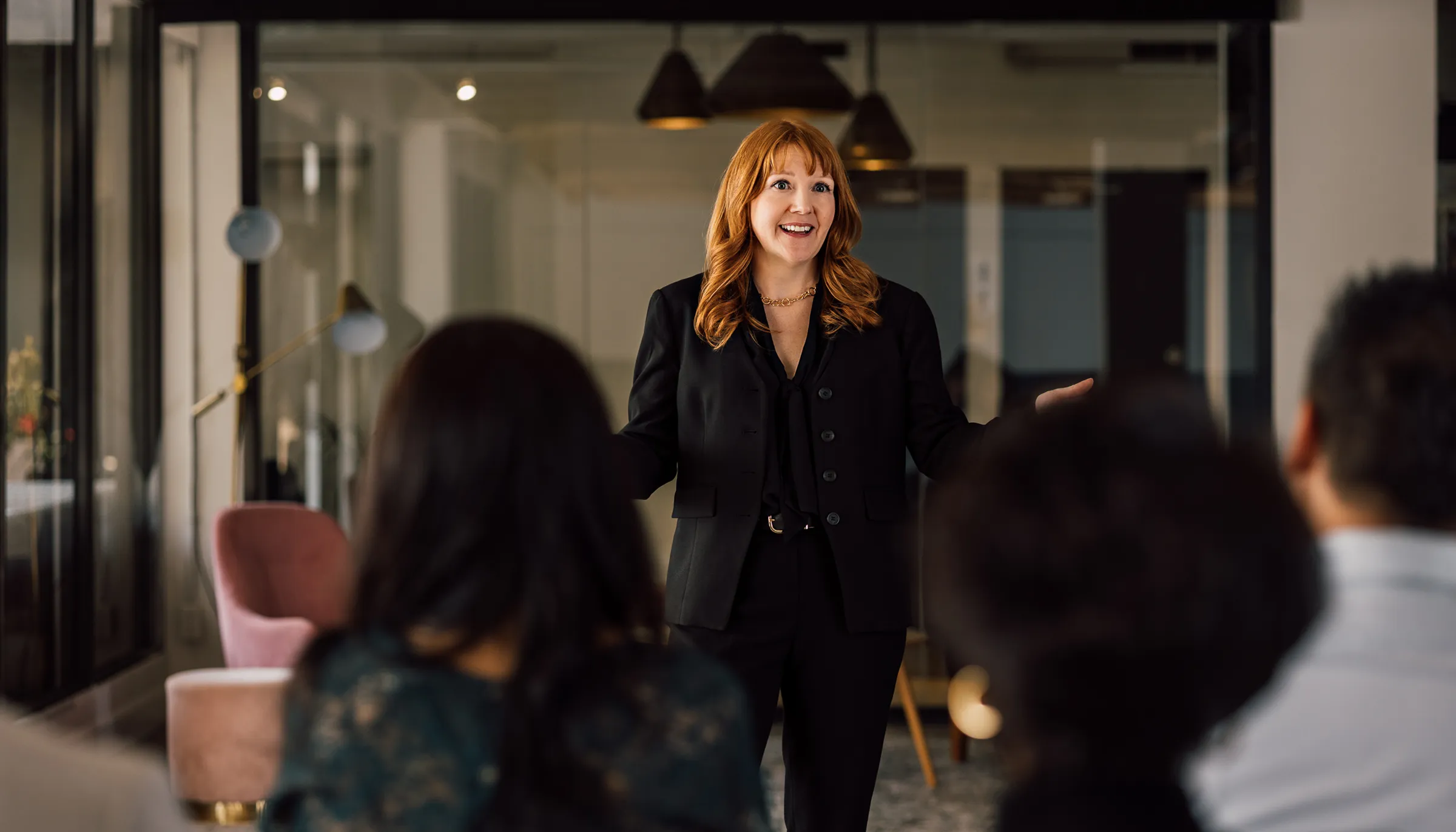 Red-haired woman in a black suit smiling and speaking confidently to an audience in a modern office setting.