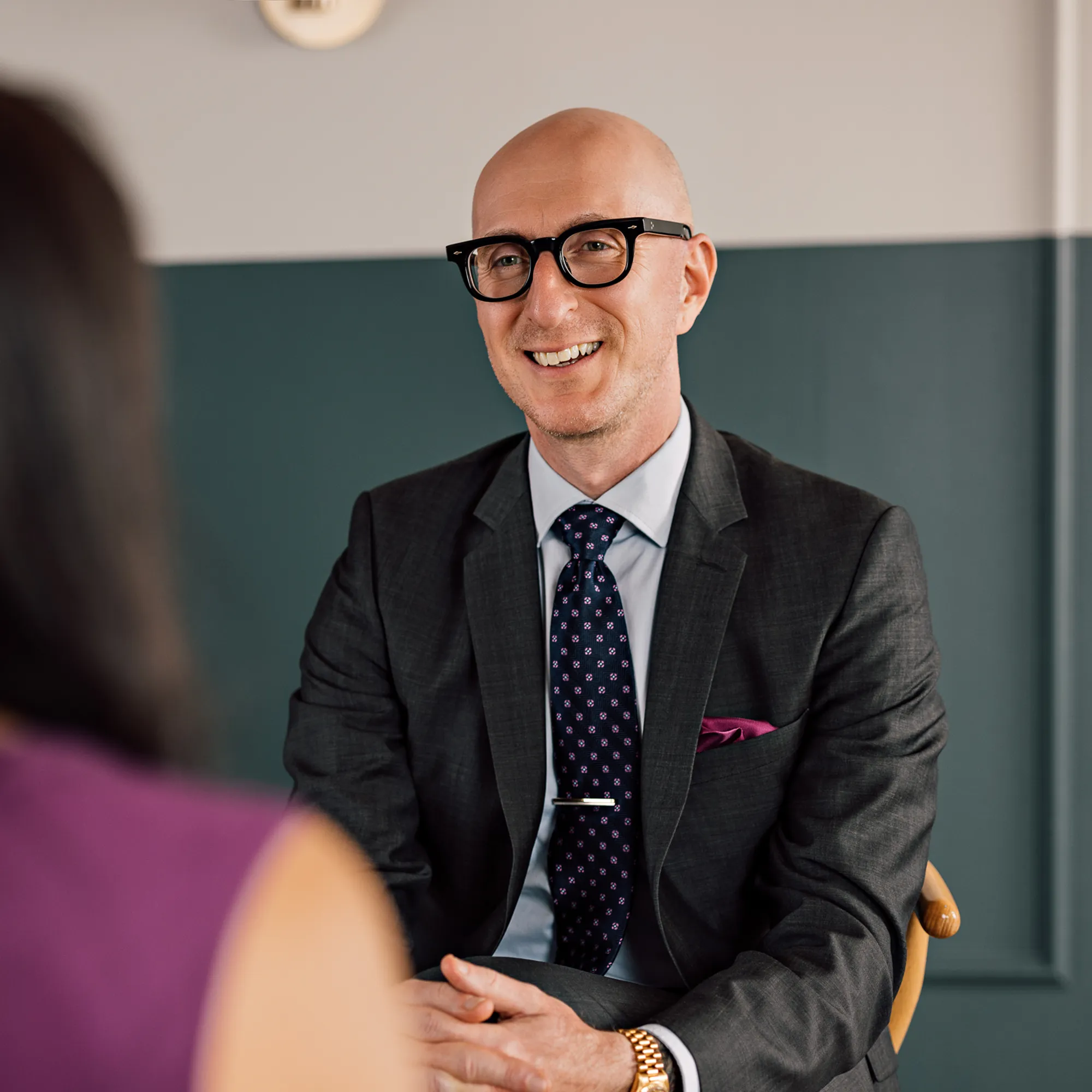 Man in glasses and a charcoal suit smiles while listening to a woman across from him during a coaching session