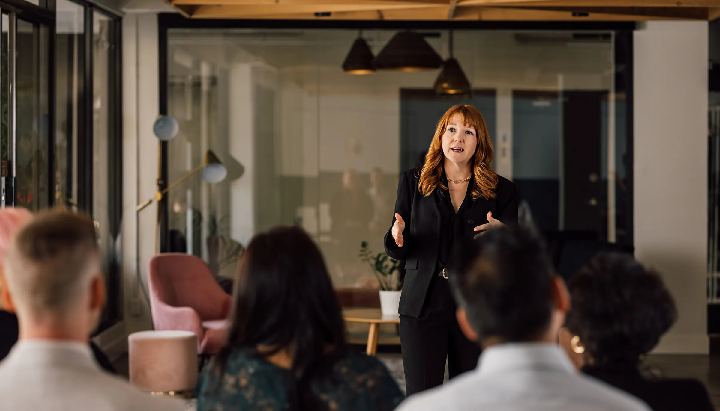 Woman with red hair wearing a black suit speaking and gesturing with her hands in front of an audience in a modern office setting.