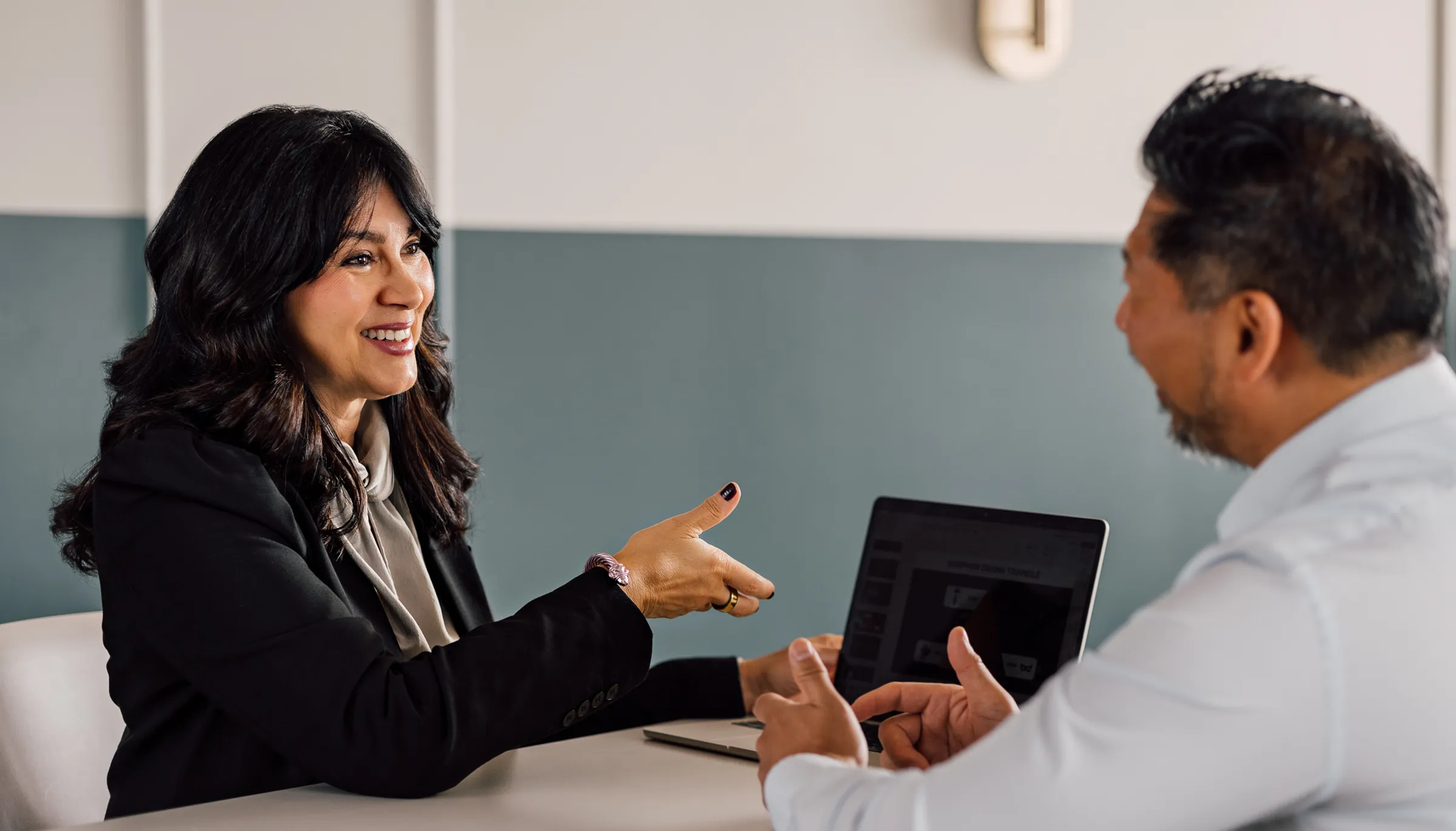 Woman in a black blazer gestures while talking with a man across a table with an open laptop