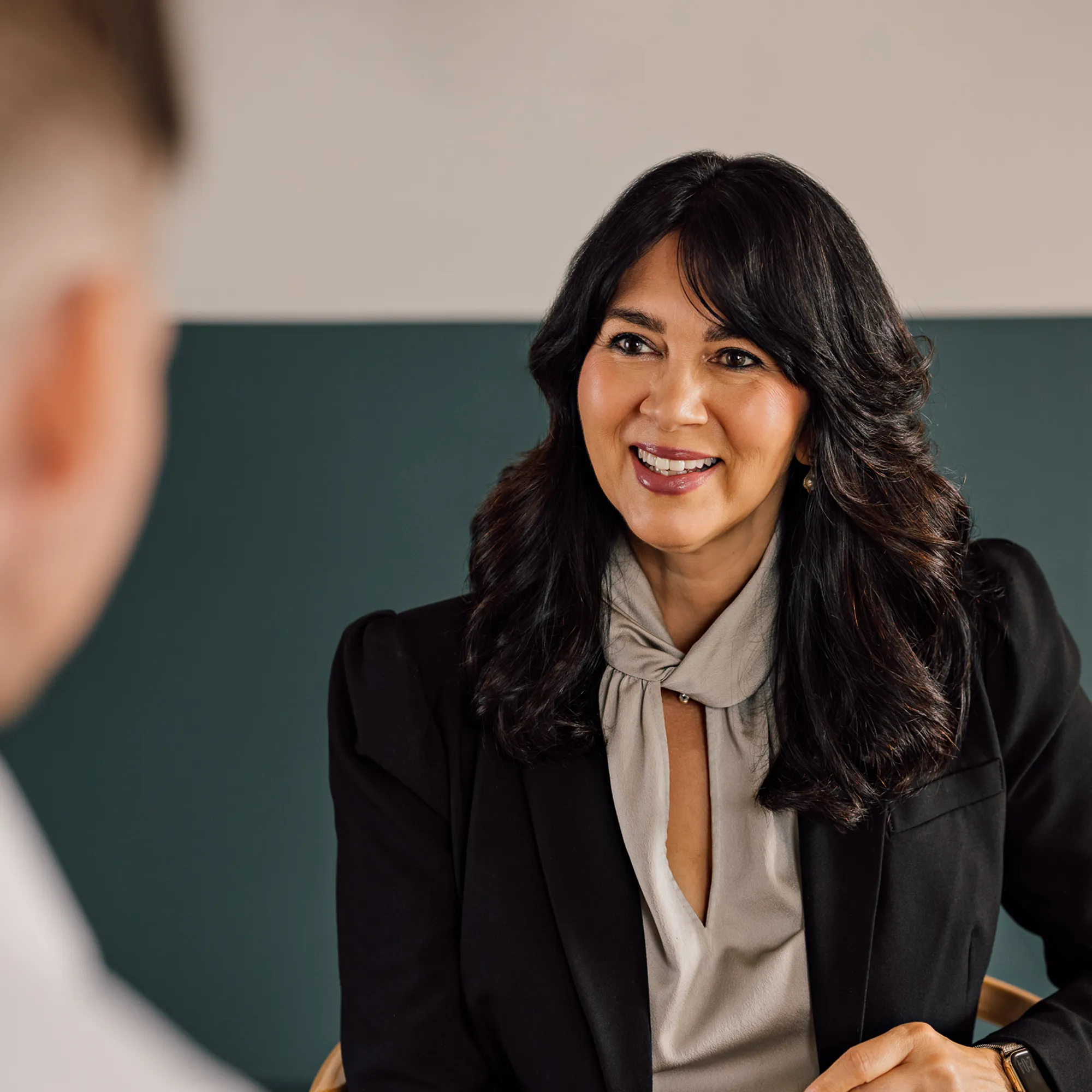 A professional woman with long dark hair in a black blazer and light blouse is smiling and conversing with a person facing away from the camera.