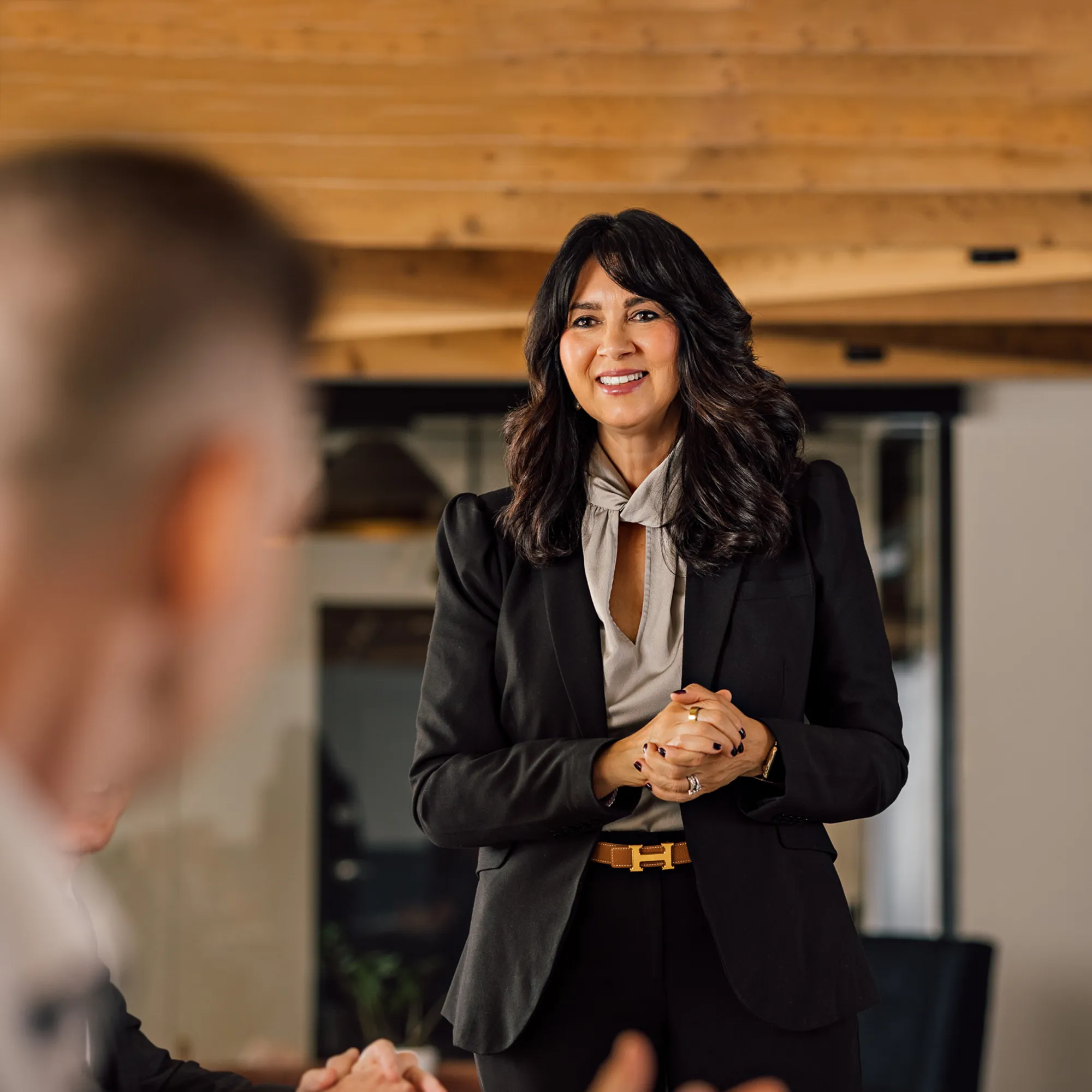 Smiling professional woman with dark wavy hair wearing a black blazer and beige blouse speaking in a modern office with wooden ceiling beams, facing blurred individuals.
