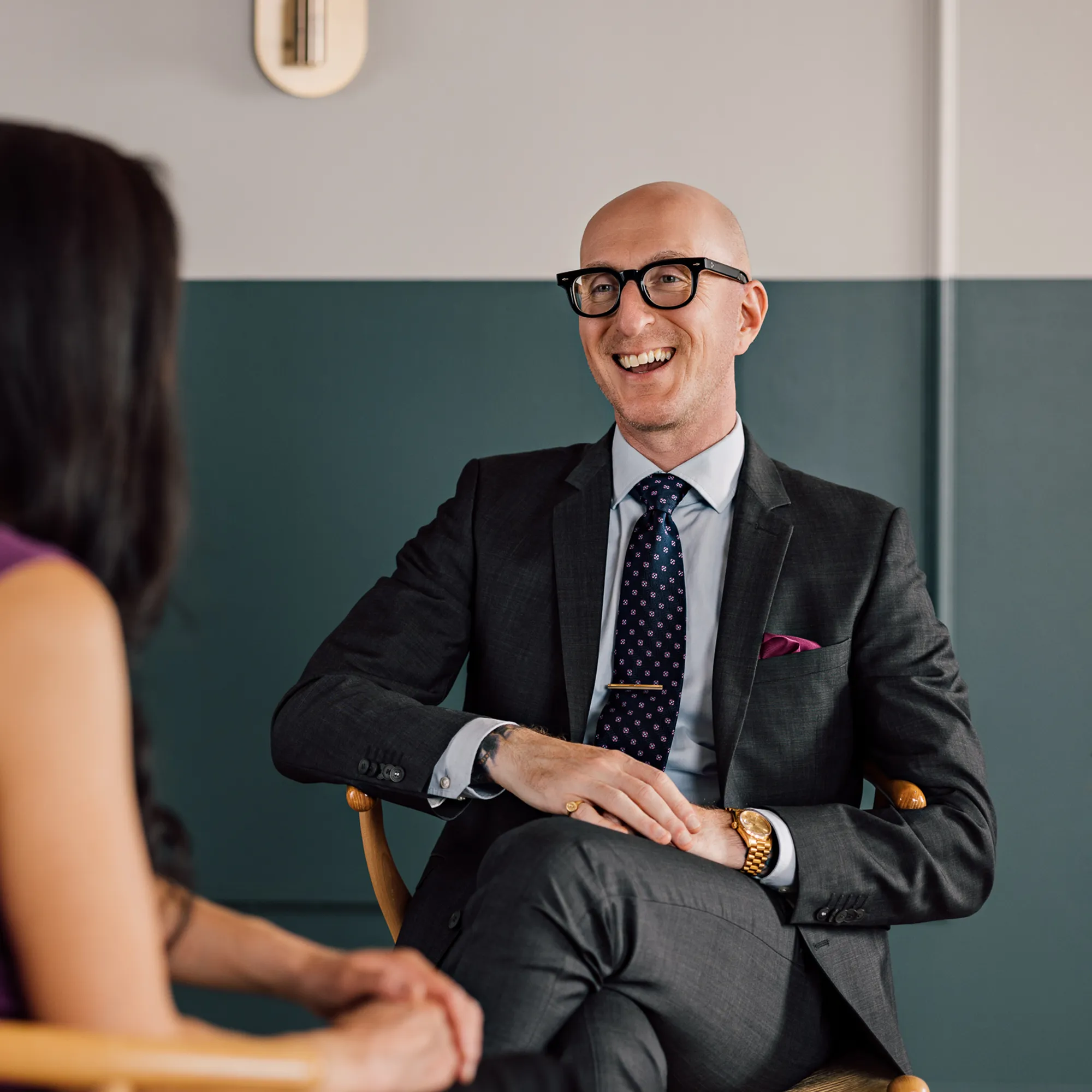 Man in glasses and a suit laughs during a relaxed conversation, seated across from a woman