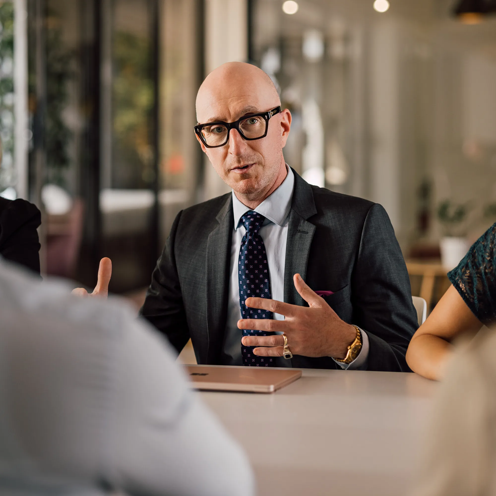 Man in glasses and a suit talks to a group of people seated at a table
