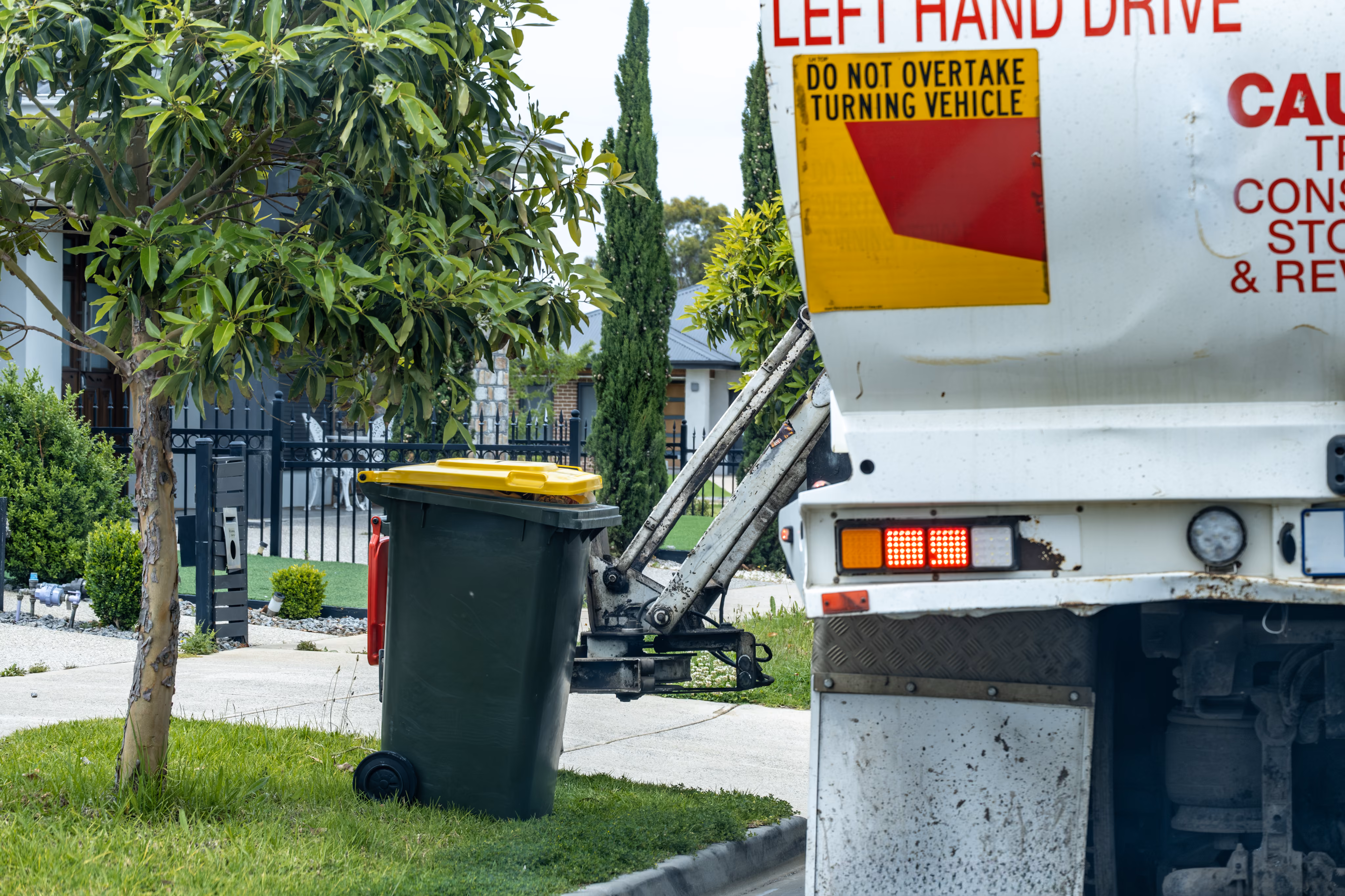 Side-lift collection arm gripping a kerbside wheelie bin beside a residential property