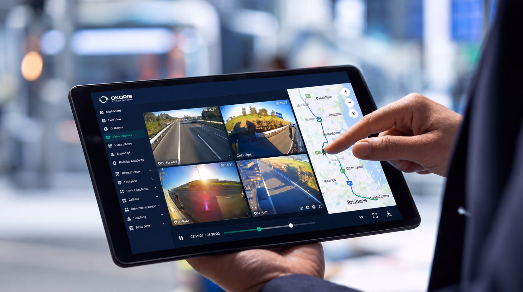 Hand holding a tablet showing OKORIS video playback with four camera channels and the vehicle route on a Brisbane map