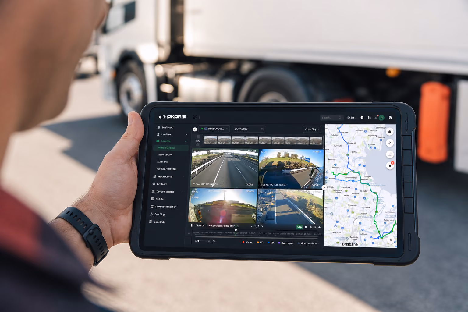 Fleet manager holding a rugged tablet showing OKORIS video playback and route mapping, beside a parked truck