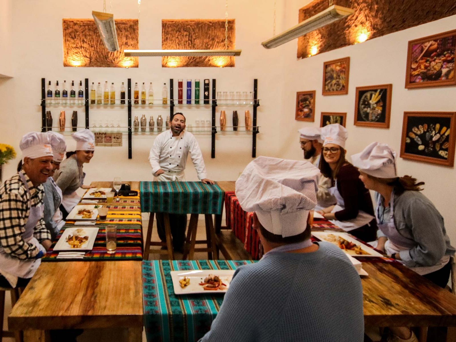 A chef in a white toque leads a lively hands-on cooking class, as guests in paper chef hats enjoy plated Peruvian dishes at a rustic wooden table.