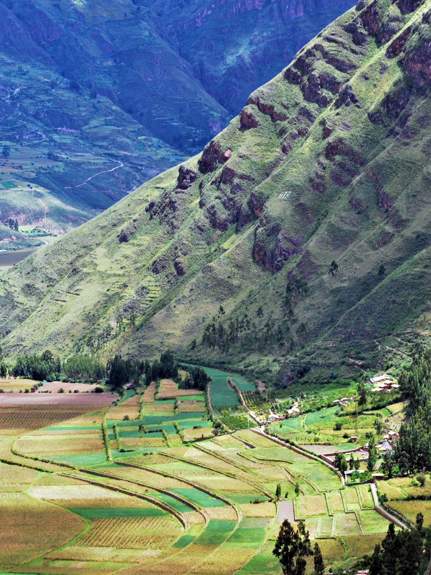 A quilt-like patchwork of green and gold farm plots lines the Sacred Valley floor, a river winding beneath steep striated mountainsides.