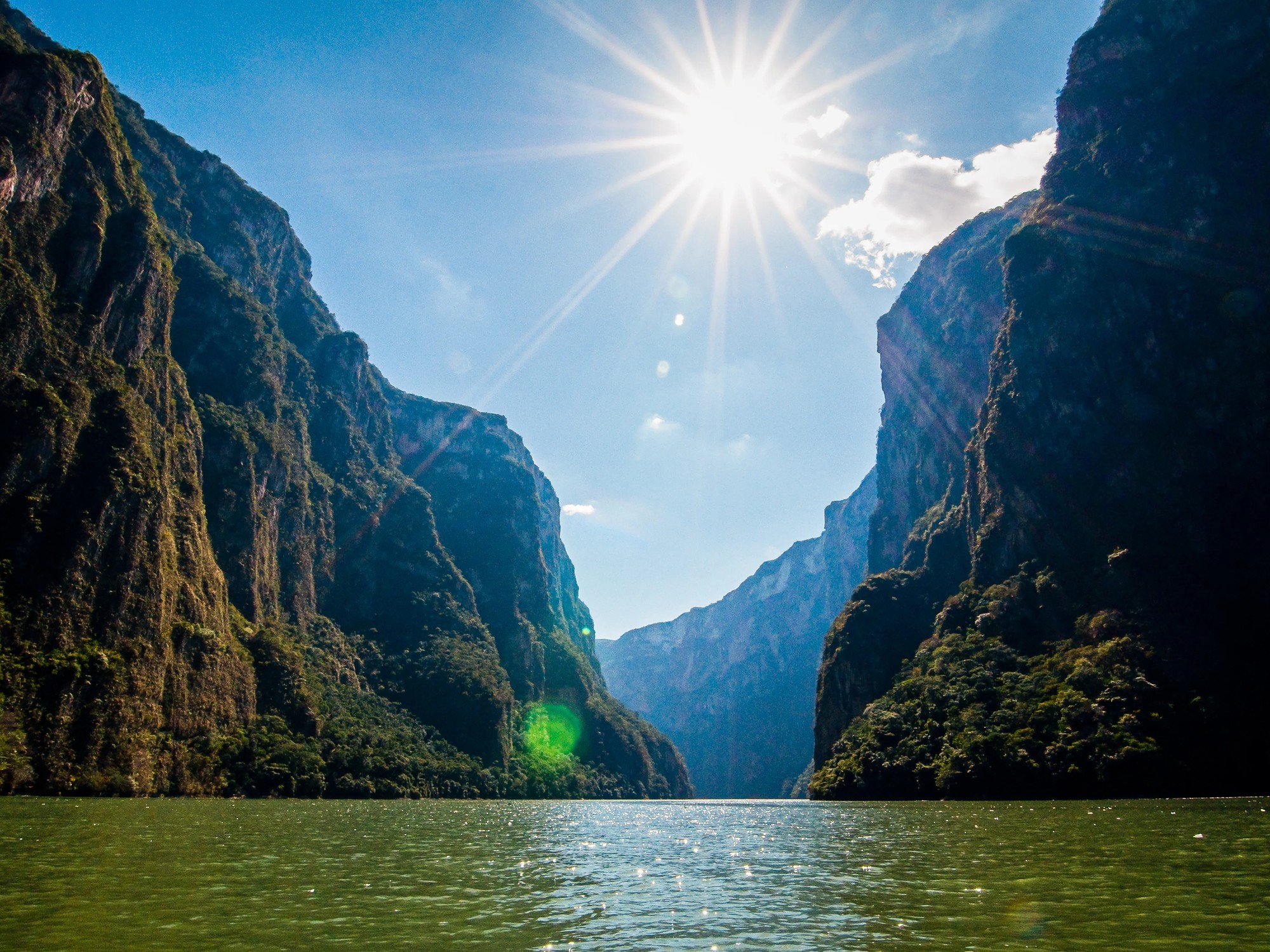 Sunlight bursts between the towering cliff walls of Sumidero Canyon above the calm green river below.
