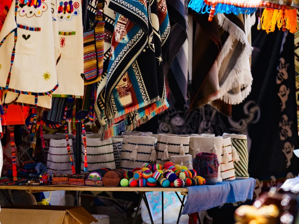 Colorful hand-woven ponchos, tasseled baskets, and knitted balls fill a sunlit stall at the Otavalo market.