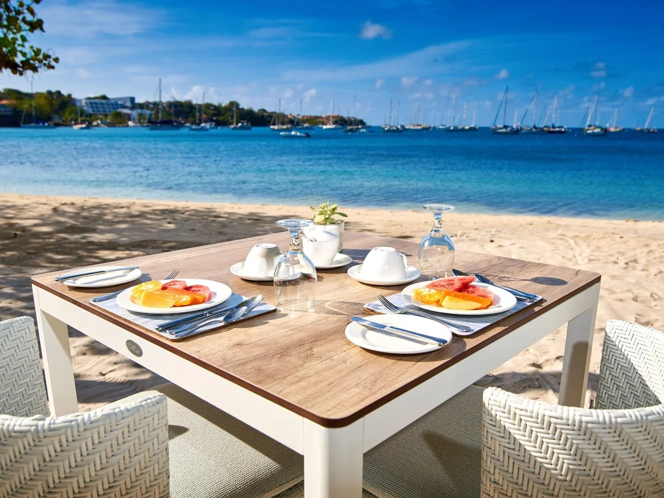Breakfast table set on a sandy beach overlooking a calm bay.