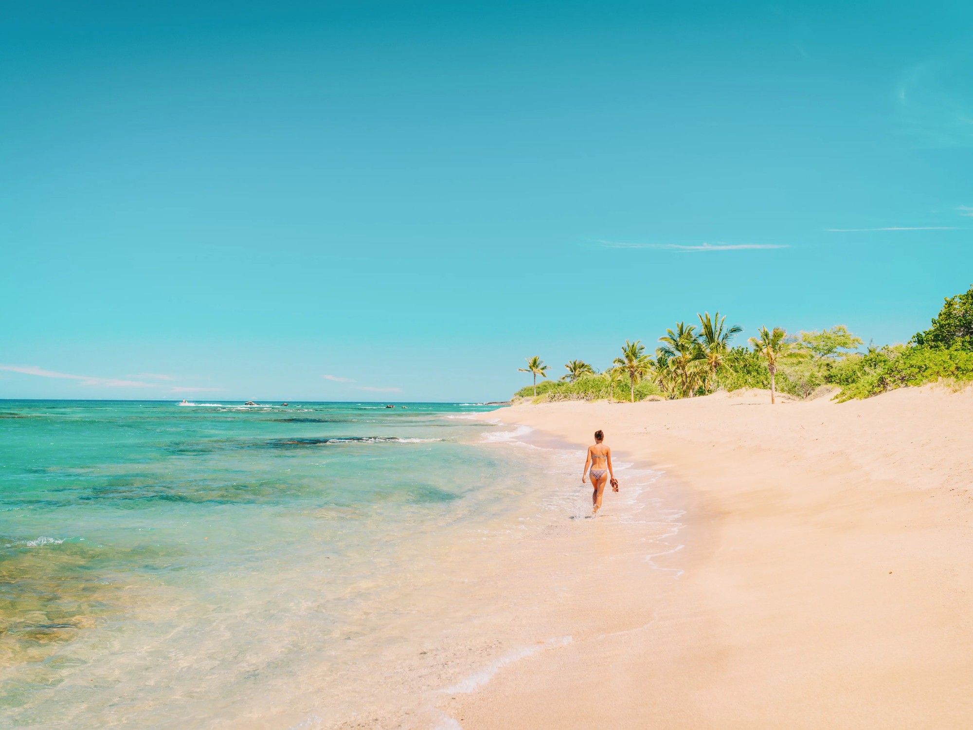 A woman in a bikini strolls barefoot along the shoreline of an empty Grenadines beach.