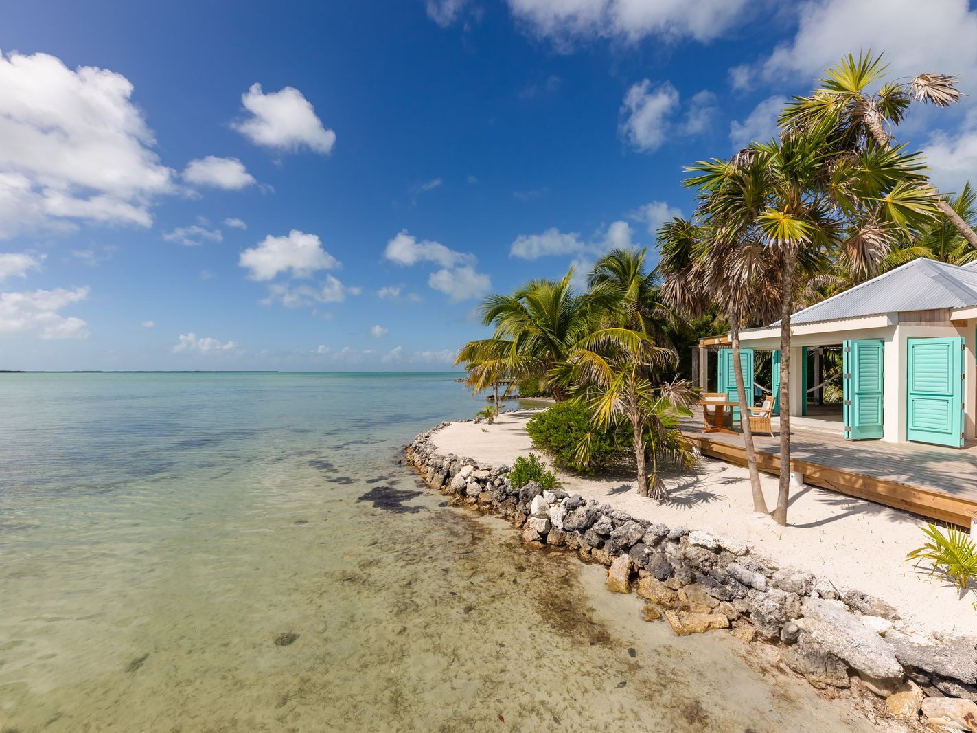 A private island beach cottage with turquoise shutters sits amid palm trees and clear water.