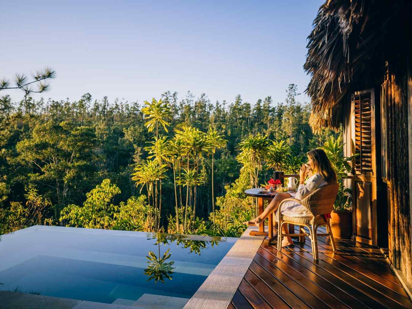 A guest enjoys breakfast on a deck beside an infinity pool overlooking the rainforest canopy.
