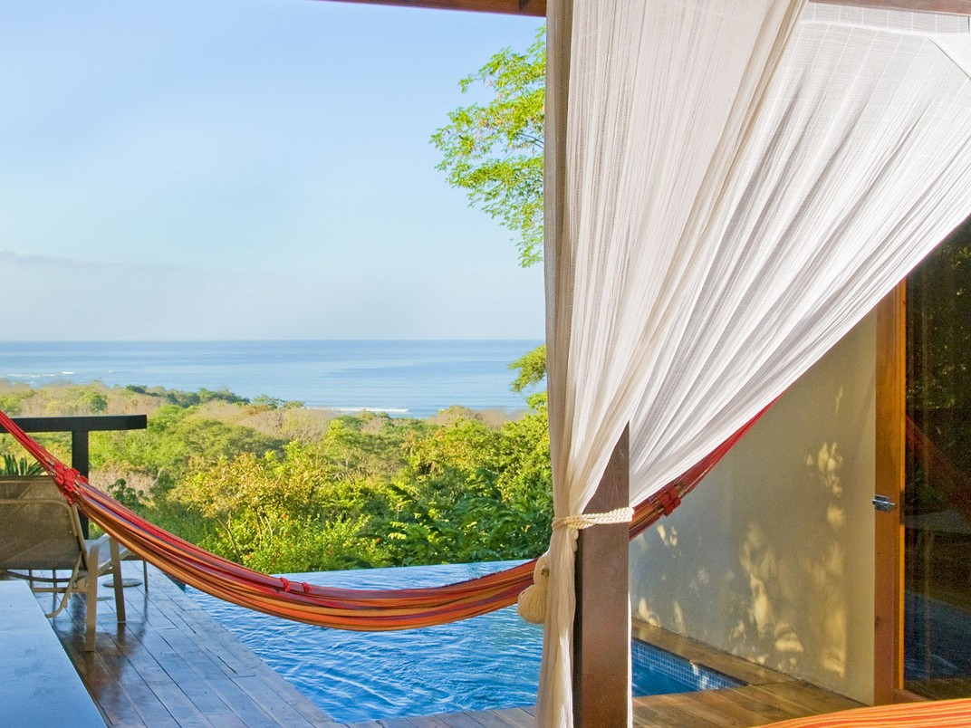 A striped hammock strung across a shaded deck beside an infinity pool overlooking the jungle canopy and Pacific coastline in Nicoya, Costa Rica.