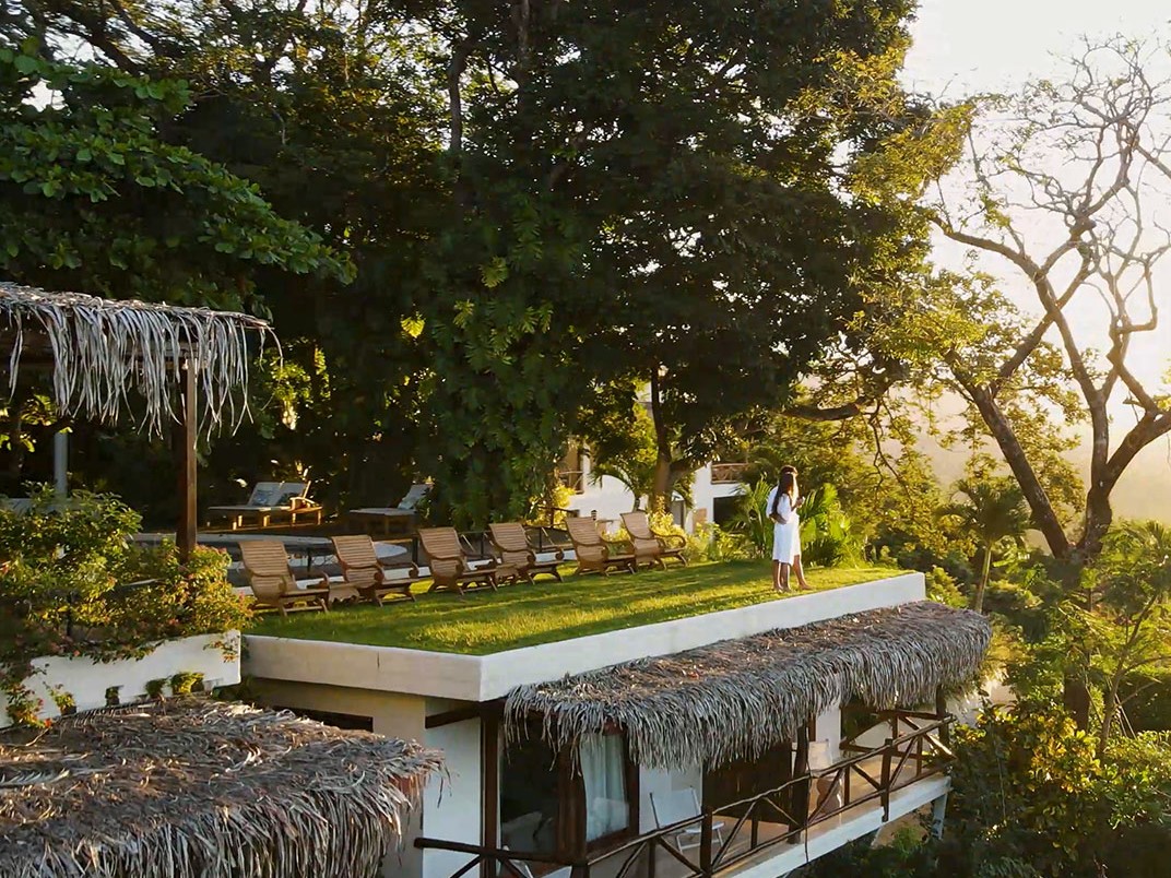 A woman in a robe stands on a grassy rooftop lounge deck lined with wooden chaise longues at a thatched-roof boutique hotel surrounded by rainforest.