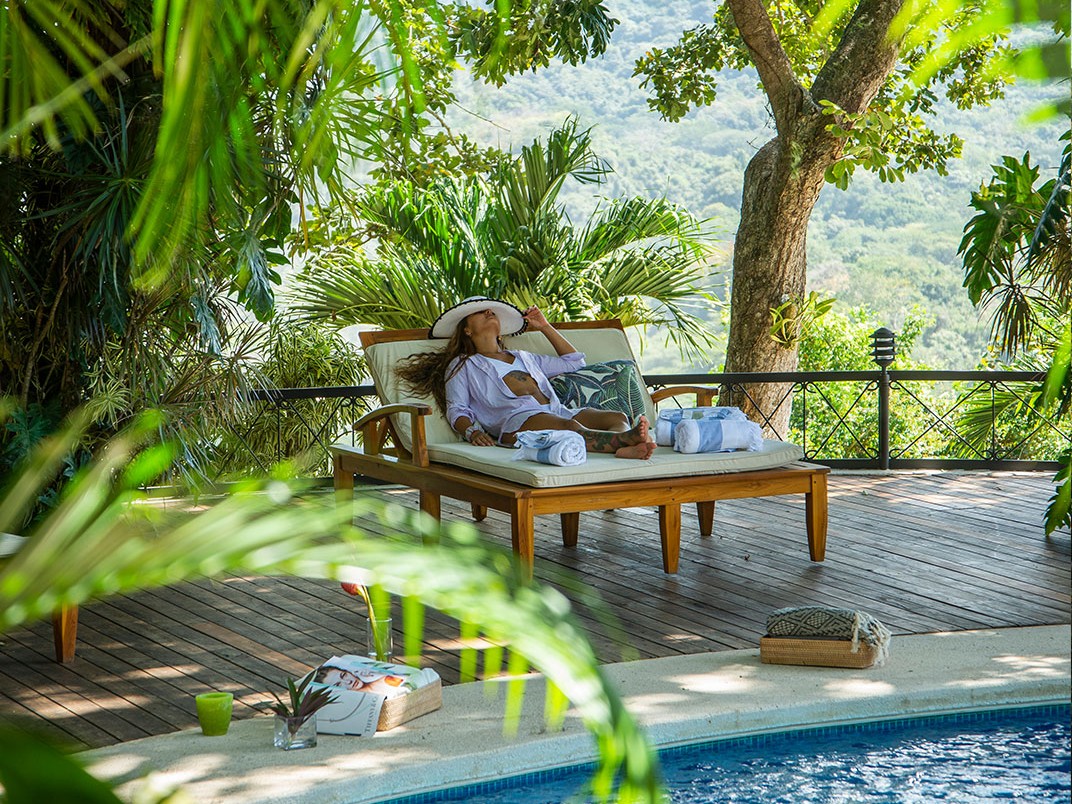 A woman in a sun hat reclines on a poolside lounge chair framed by palm fronds, with rolling jungle hills visible beyond the pool's edge.