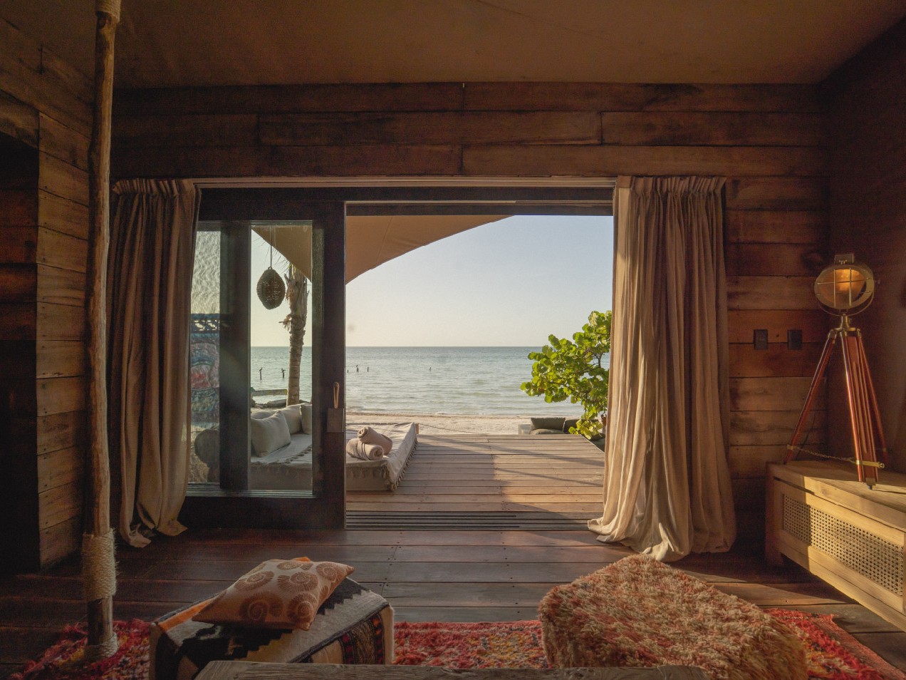 A boho-chic beachfront suite opens through wide timber doors onto a private deck and daybed, framing an unobstructed ocean view.