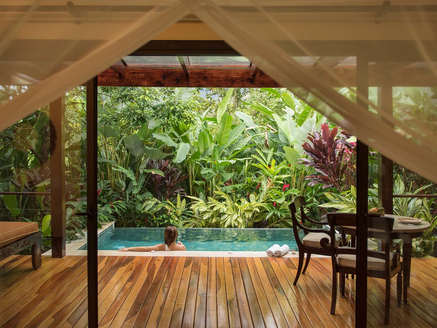 Sheer curtains frame a private plunge pool where a woman relaxes surrounded by lush tropical foliage.