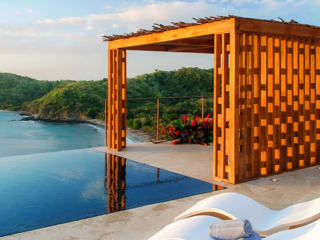 A private infinity pool terrace overlooks the ocean beneath a wooden pergola, with rolled towels on curved lounge chairs.