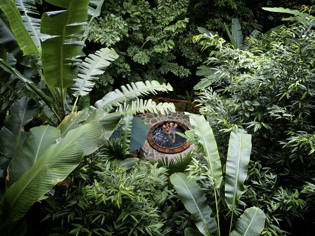 A couple soaks in a stone hot spring jacuzzi hidden among banana leaves and dense jungle foliage.