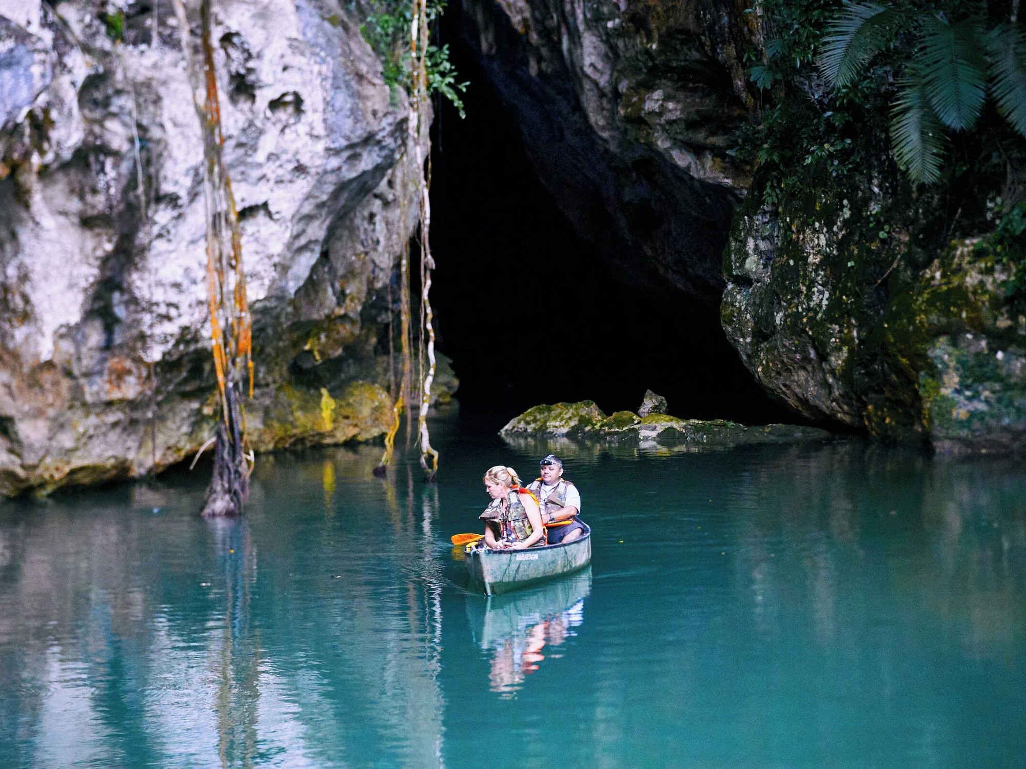Two visitors canoe toward the glowing turquoise entrance of Barton Creek Cave in Belize.