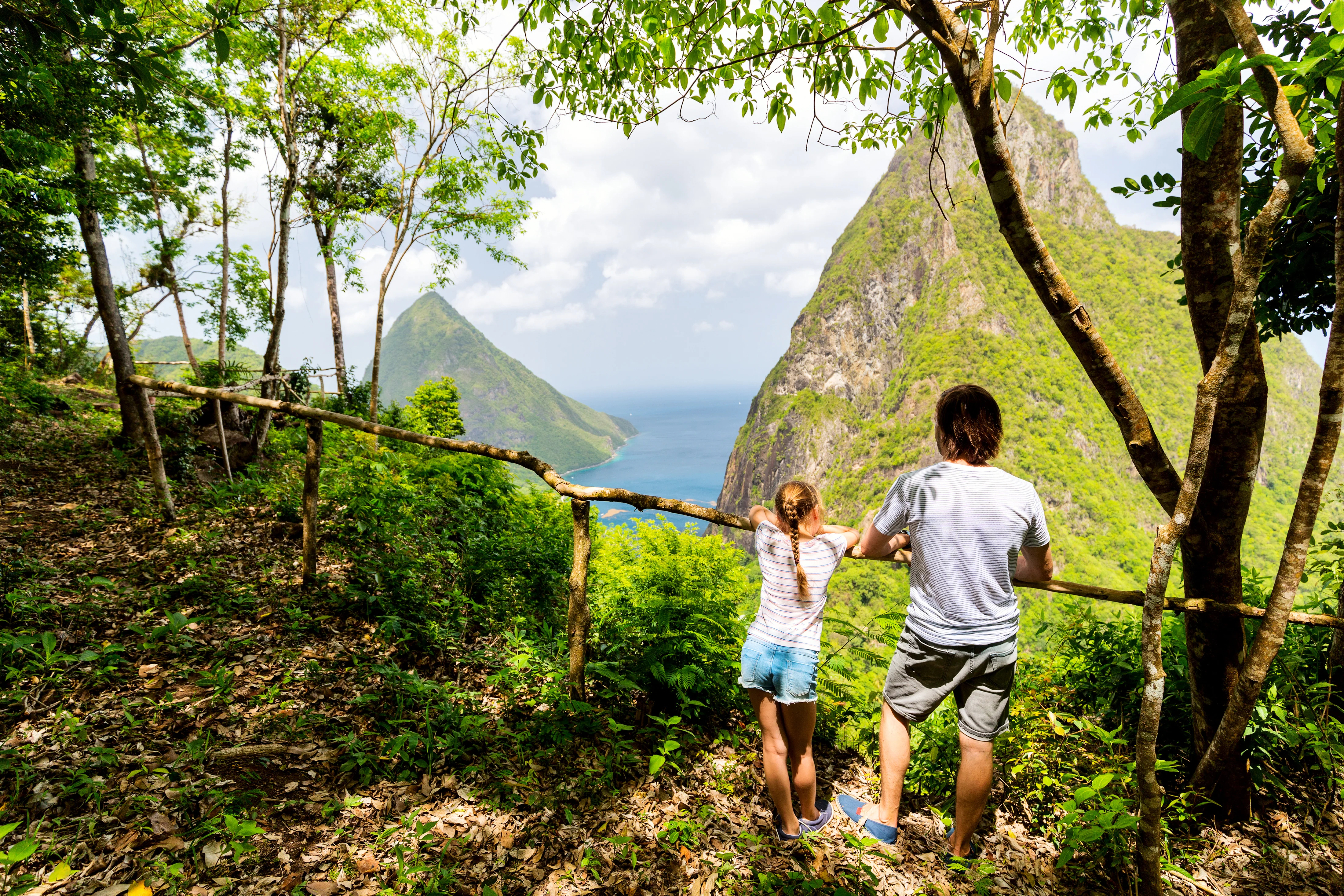 Man and girl in shorts looking at lush mountains and ocean from a forest viewpoint. St. Lucia rainforest hike, Caribbean.