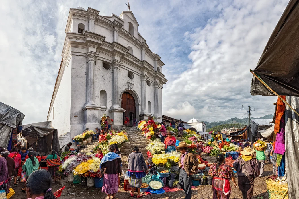 Busy market scene with vendors selling colorful flowers by stone stairs of a white colonial church. Guatemala Chichicastenango market.