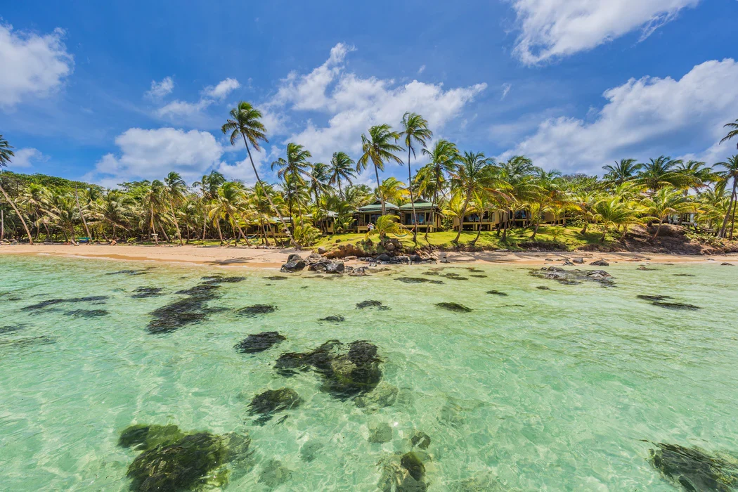 Clear turquoise water and rocky patches at a beach lined with palm trees under a blue sky with clouds. Little Corn Island, Nicaragua.