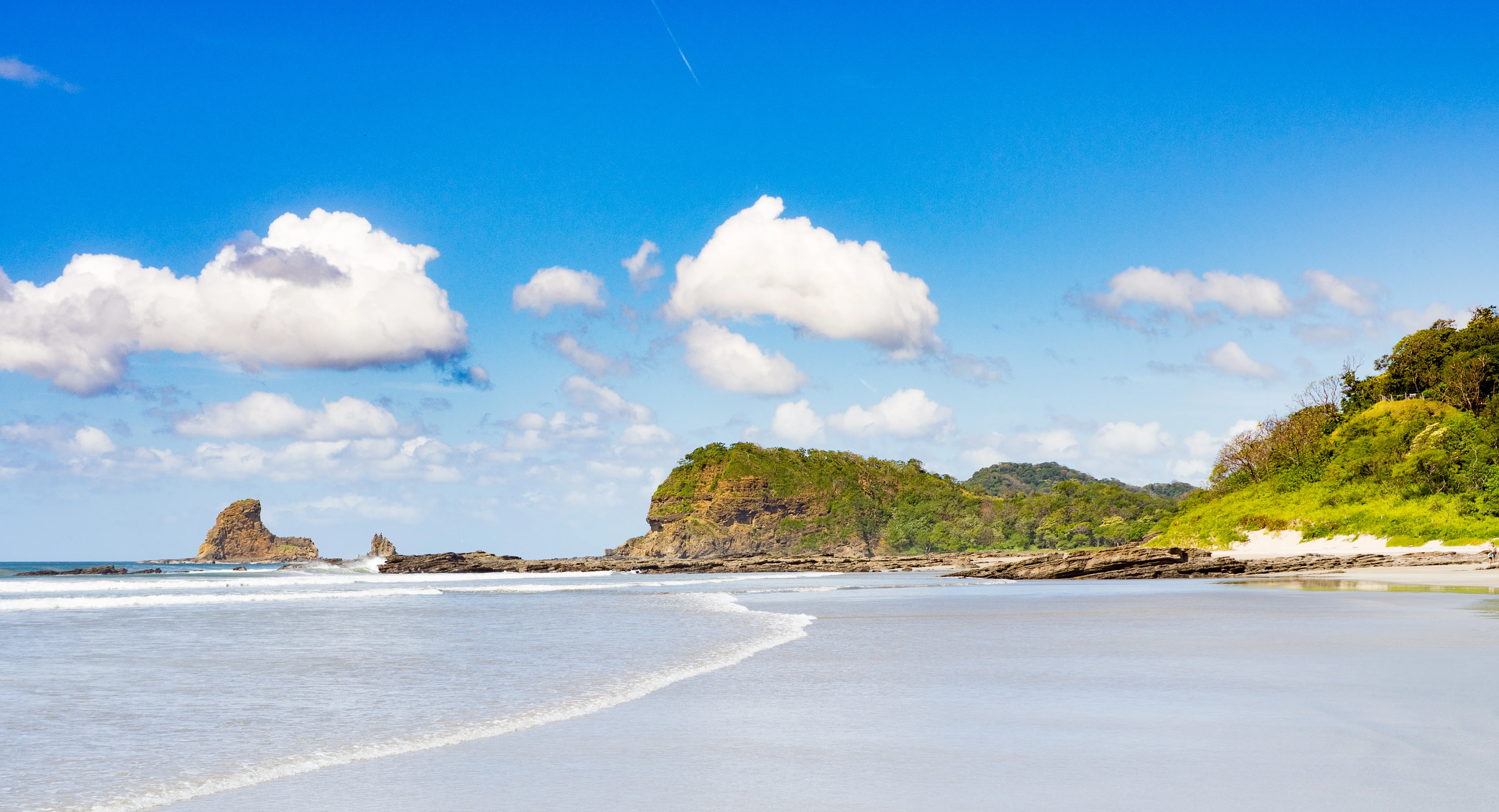 Sandy beach with gentle ocean waves, rocky cliffs, greenery, and a blue sky with scattered clouds. Playa Maderas, Nicaragua.