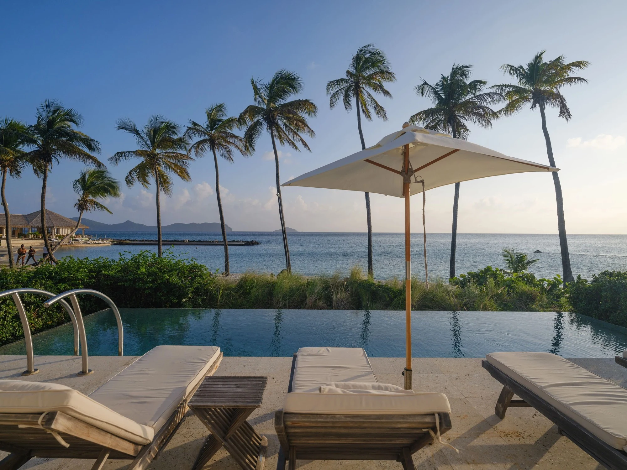 Lounge chairs and an umbrella line an infinity pool overlooking the ocean and palm trees at The Liming resort.