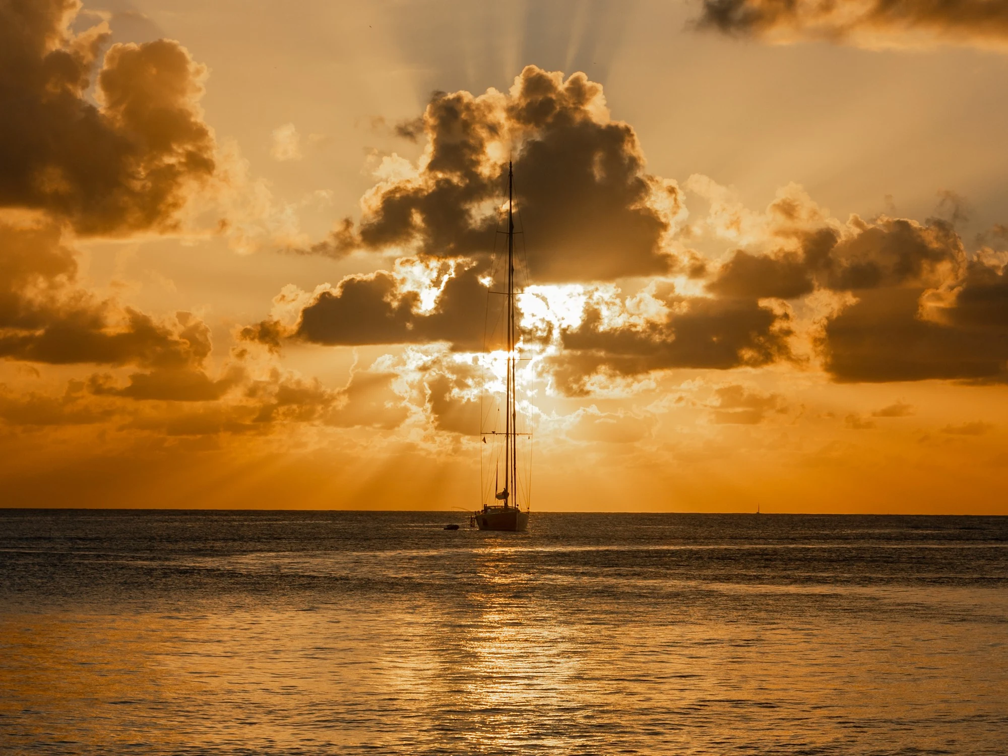 A lone sailboat silhouetted against a fiery orange Grenadines sunset with sunbeams breaking through clouds.