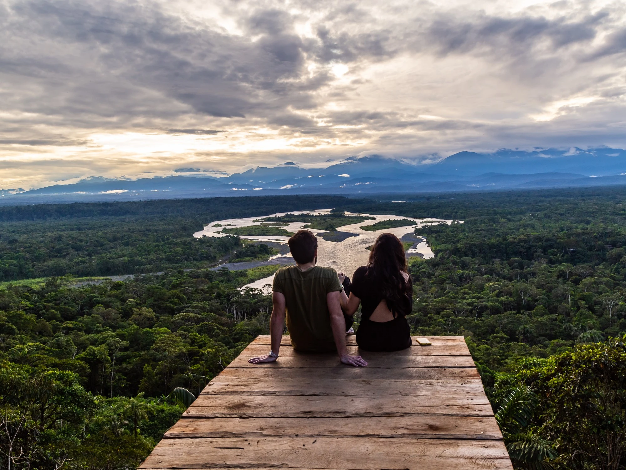 A couple sits at the edge of a wooden viewing platform, gazing out over a serpentine Amazon river bend and misty blue mountains at dusk.
