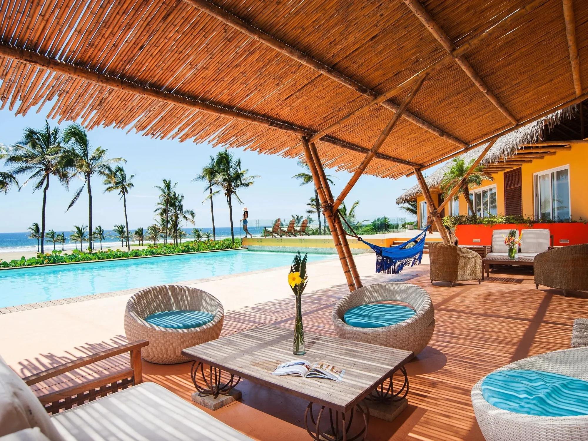 Woven lounge chairs and a blue hammock line an infinity pool deck beneath a thatched palapa roof, with palm-fringed ocean stretching beyond.