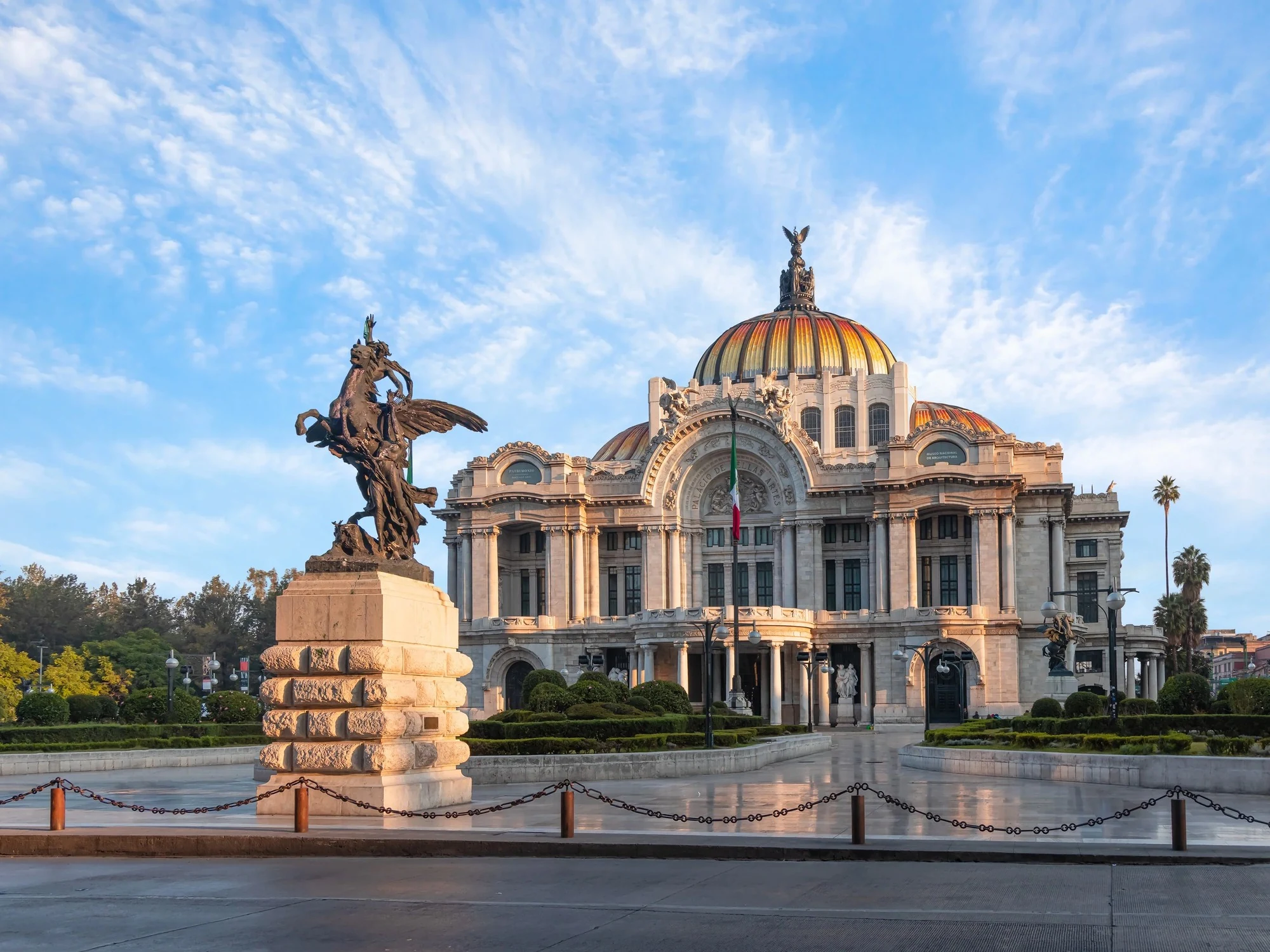 The white marble facade and golden dome of the Palacio de Bellas Artes glow in early morning light beside its bronze Pegasus monument.