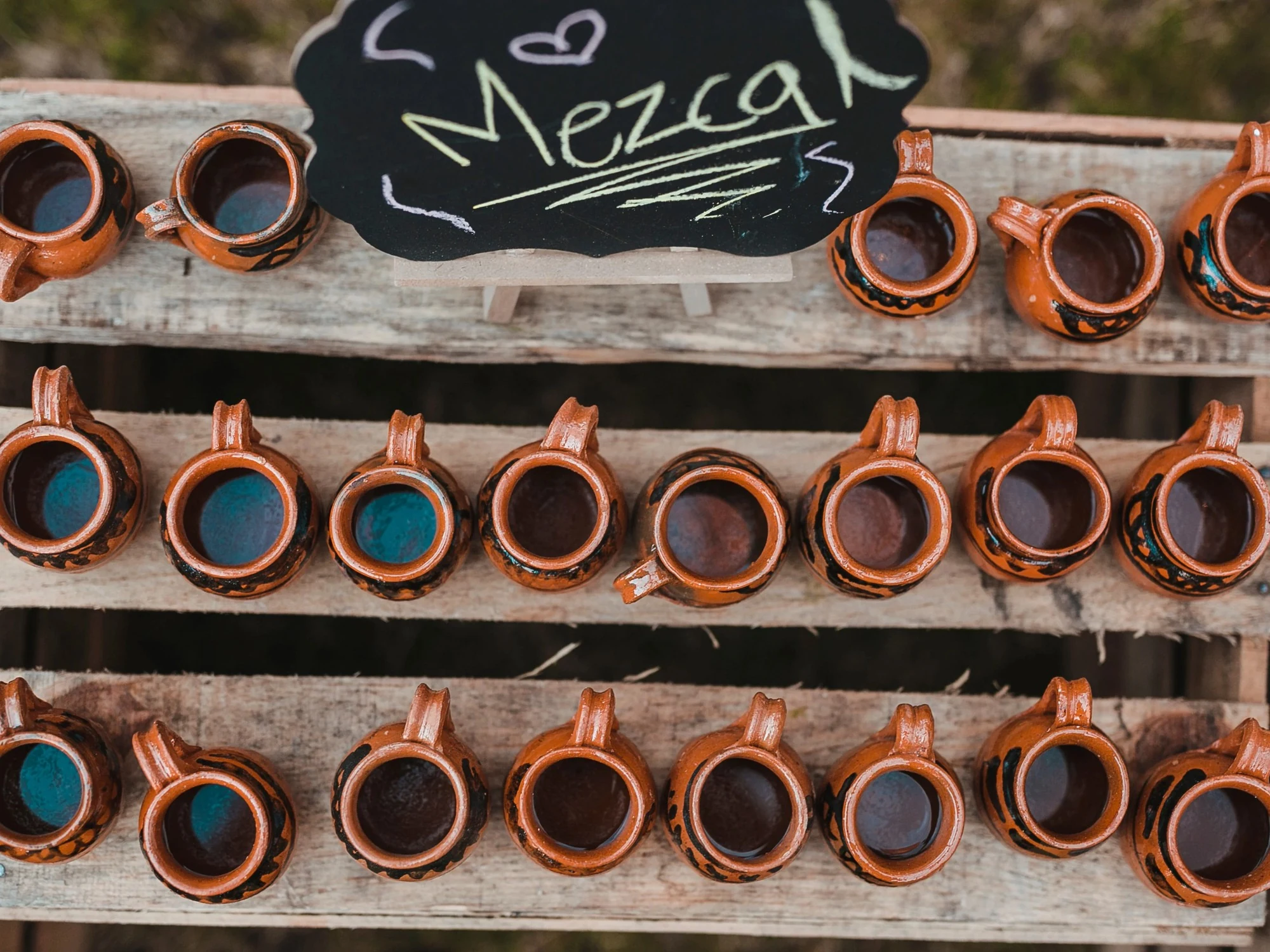 Rows of small clay cups line a wooden mezcal tasting stand marked with a handwritten chalkboard sign.