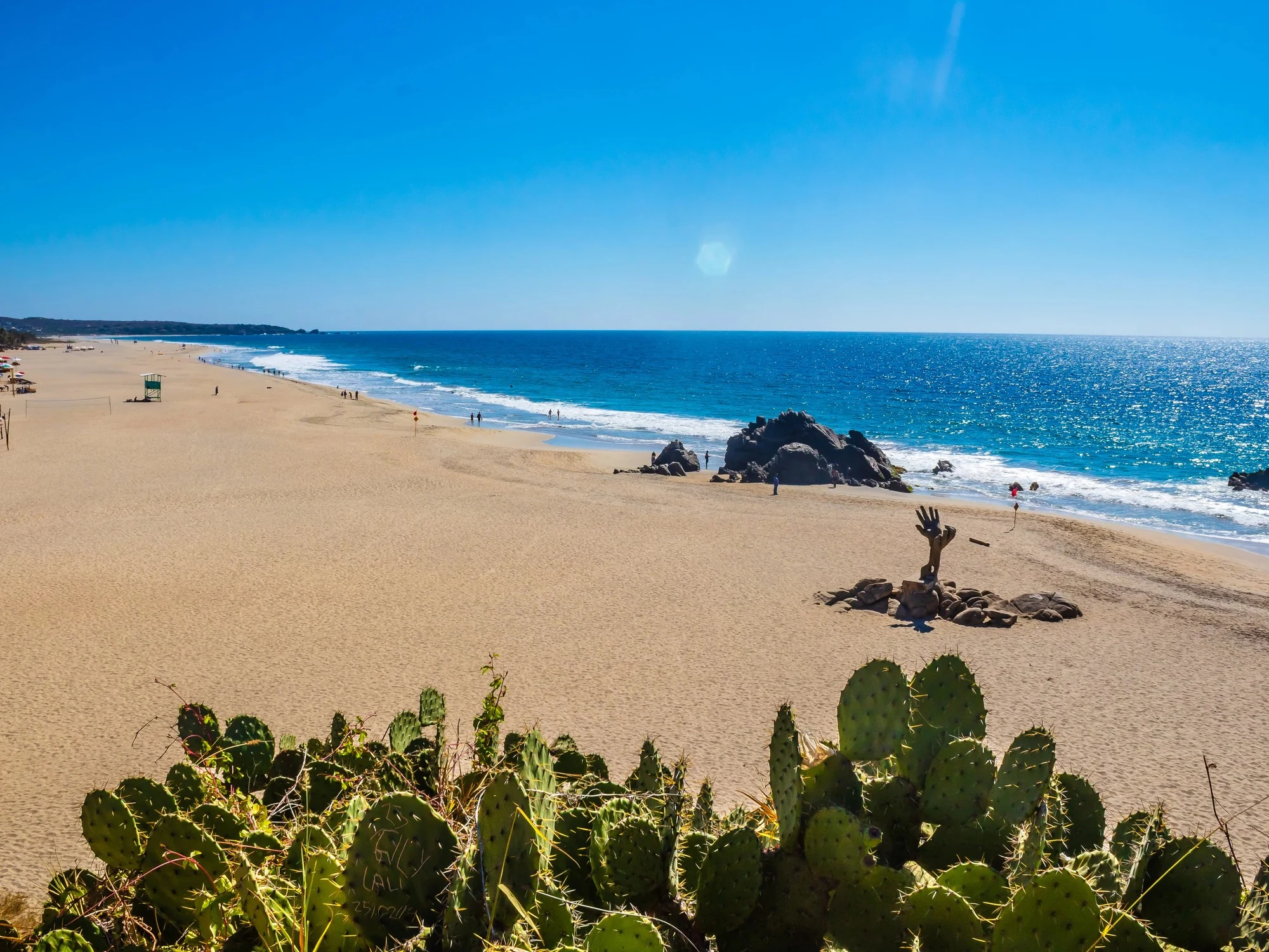 A giant hand sculpture rises from the sand on Zicatela Beach, framed by cactus and volcanic rock in Puerto Escondido.