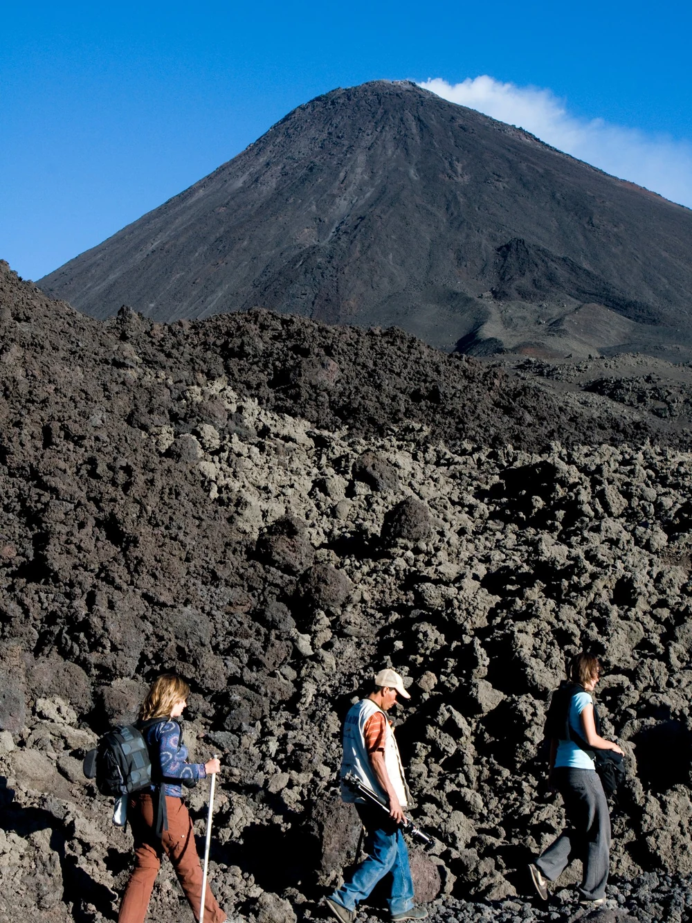 Hikers cross a black lava field with a smoking volcanic cone rising behind them.