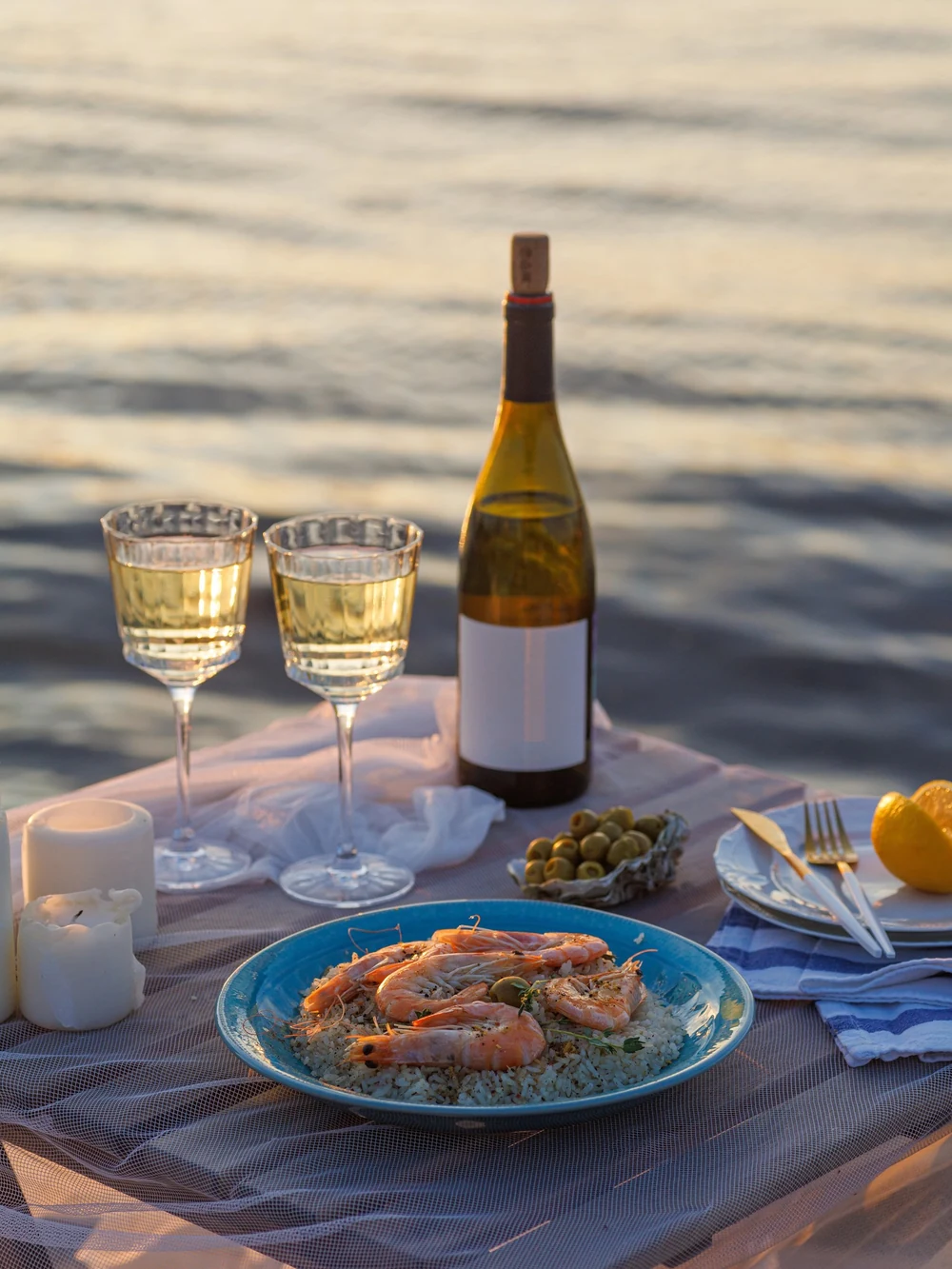 A candlelit beachfront table set with white wine, olives, and grilled shrimp overlooks the shimmering sea at dusk.