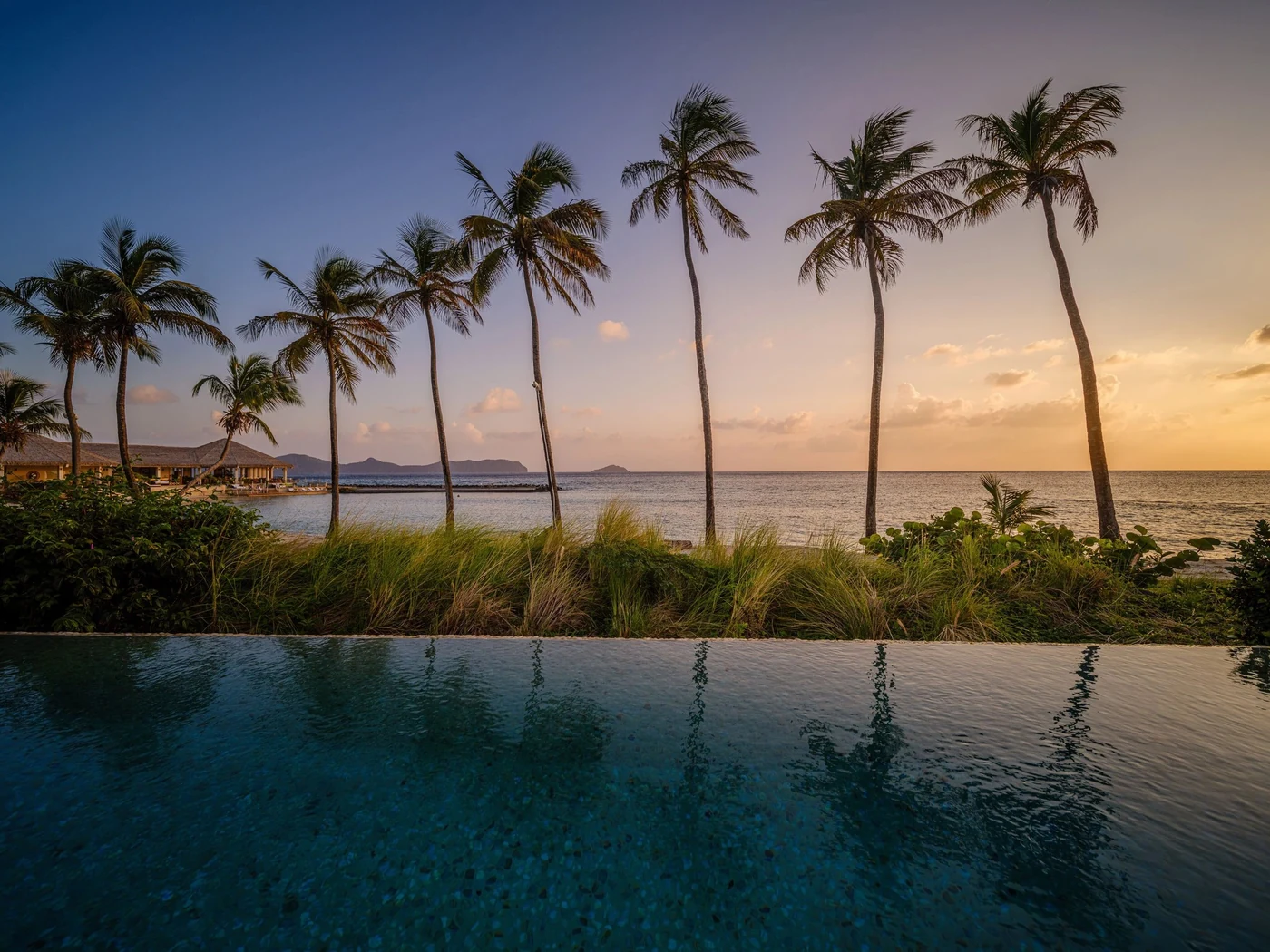 Tall palm trees silhouette against a pastel sunset sky beside the infinity pool at The Liming resort.